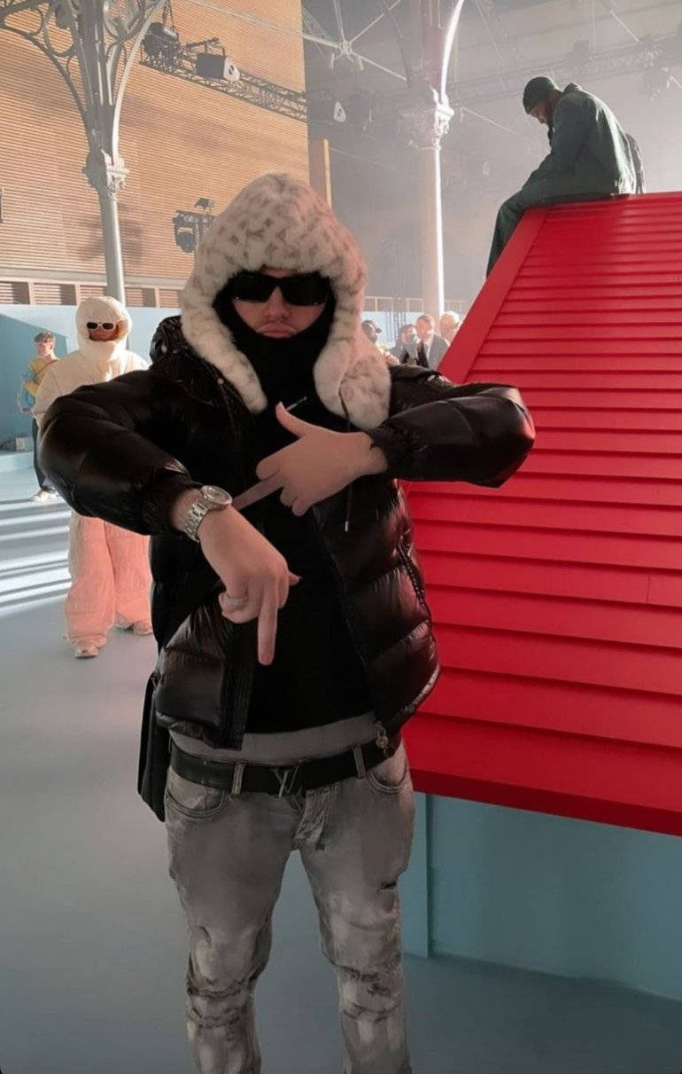 Celebrity 桌布 壁紙 — 一名身穿黑色羽絨外套和毛絨連帽的男子站在現代室內環境中，指著手錶，旁邊是鮮紅色的樓梯。