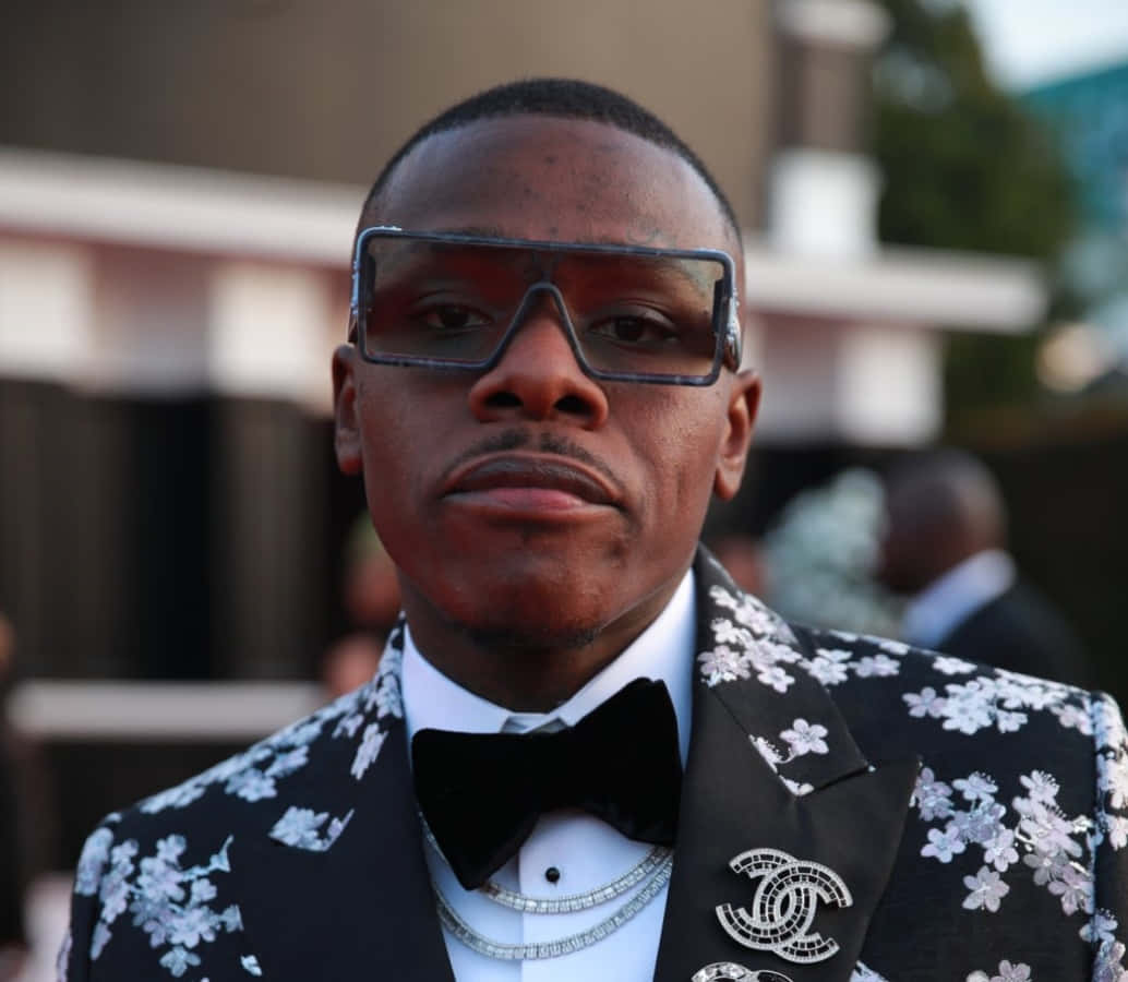 Dababy 桌布 壁紙 — 一名身穿黑色花卉西裝和超大眼鏡的男子在活動中自信地擺姿勢，背景模糊。