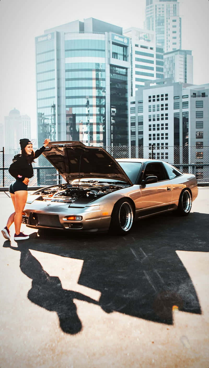 Nissan 180sx 桌布 壁紙 — 一名身穿黑色衣服的女子站在一輛打開引擎蓋的銀色跑車旁邊，背景是高樓大廈的屋頂停車場。