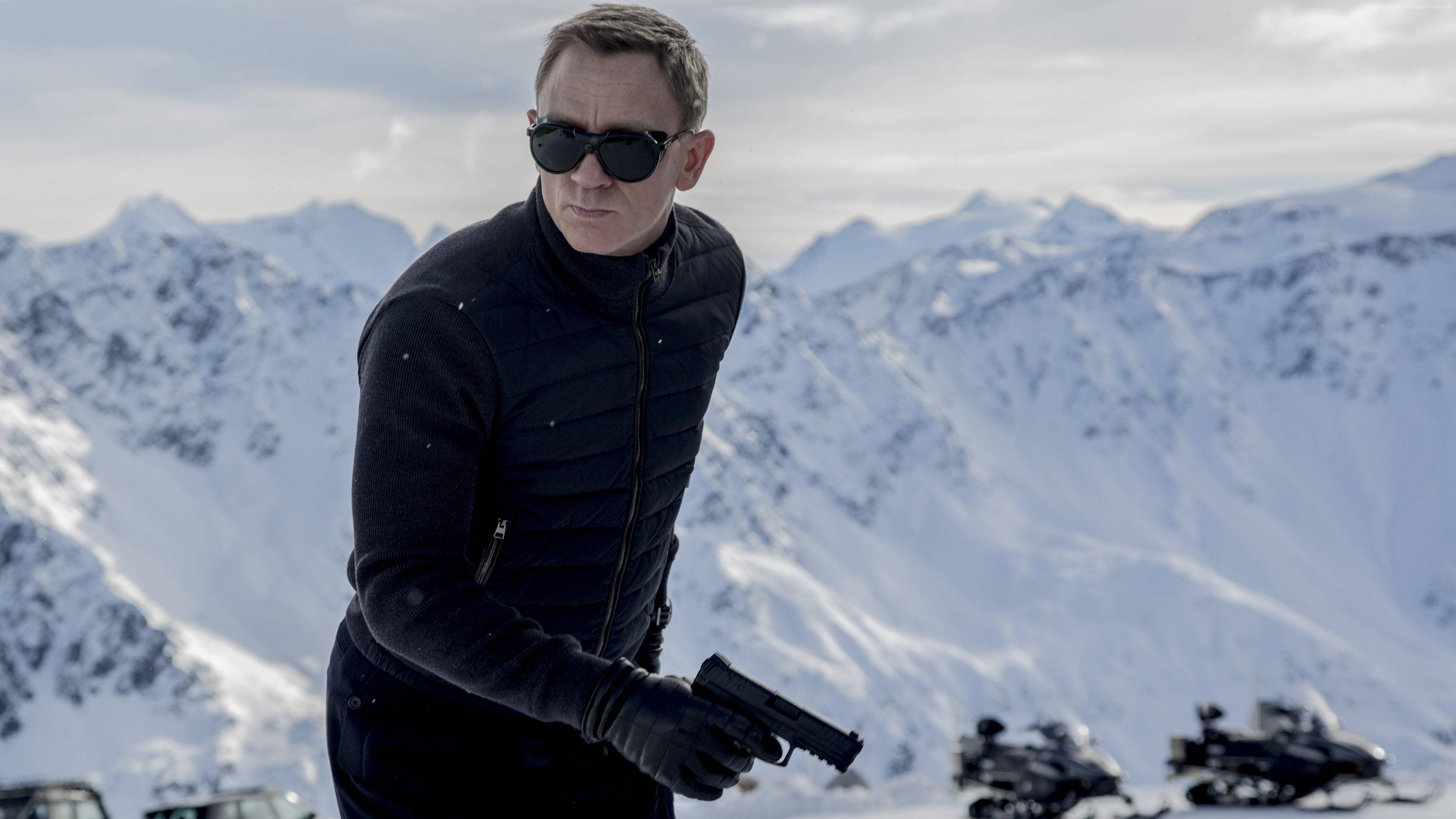 Daniel Craig 桌布 壁紙 — 一名身穿黑色裝備、戴著太陽眼鏡的男子，在陰天背景下，以積雪覆蓋的山脈為背景，手持一把槍。