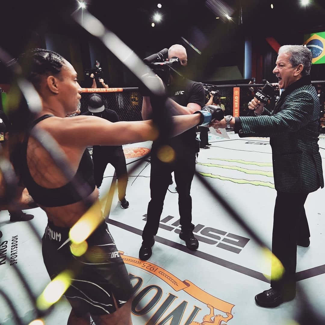 UFC 桌布 壁紙 — 一名身穿黑色裝備的女戰士在UFC八角籠中，在模糊的燈光和攝影機下向評論員揮拳。