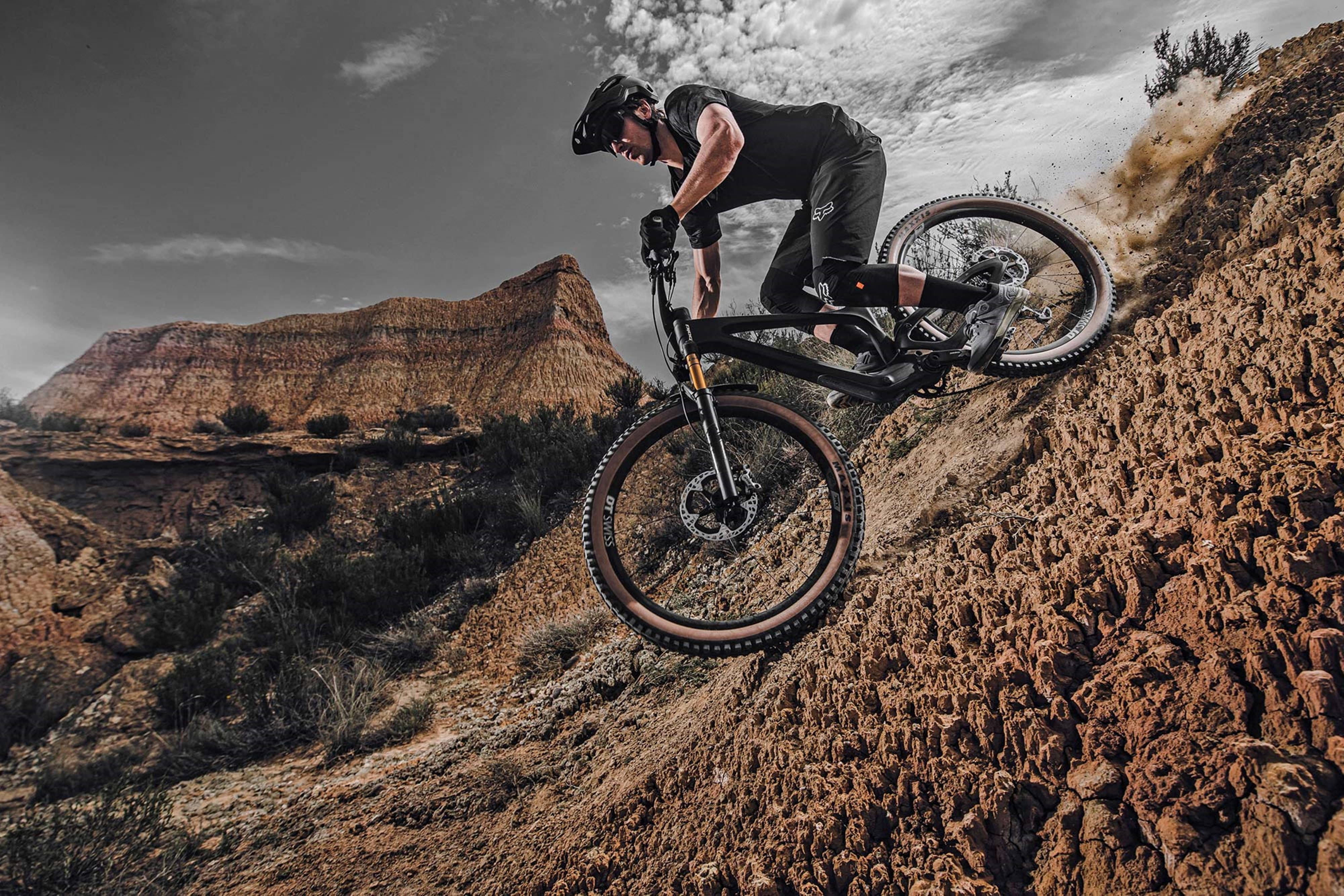 Mountain Bike 桌布 壁紙 — 一名身穿黑色裝備的山地車手在崎嶇的泥徑上騰空躍起，背景是壯麗的峽谷。