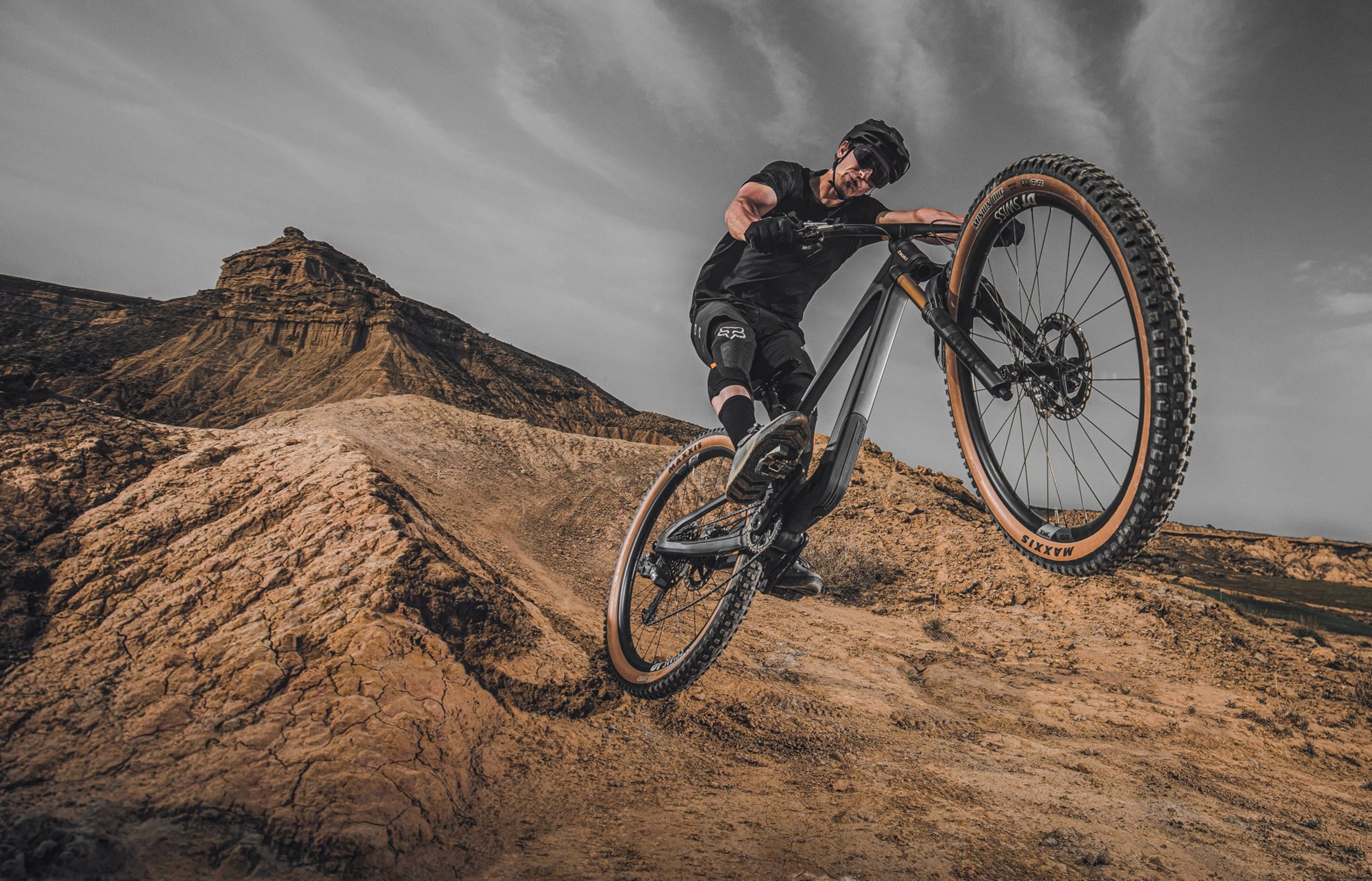 Mountain Bike 桌布 壁紙 — 一名身穿黑色裝備的山地車手在戲劇性的多雲天空下，於崎嶇的泥路上表演大膽的後輪平衡特技。