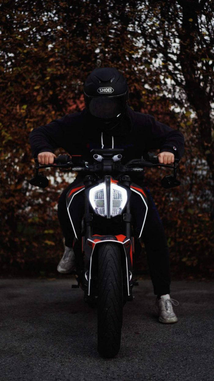 ktm rc 200 桌布 壁紙 — 一名身穿黑色裝備的機車騎士，騎著紅黑相間的機車穿過秋天的樹林，車頭燈閃閃發光。