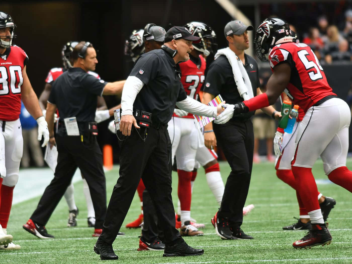 Atlanta Falcons 桌布 壁紙 — 一名身穿黑色裝備的足球教練在比賽中向綠色球場上的球員生動地做手勢。
