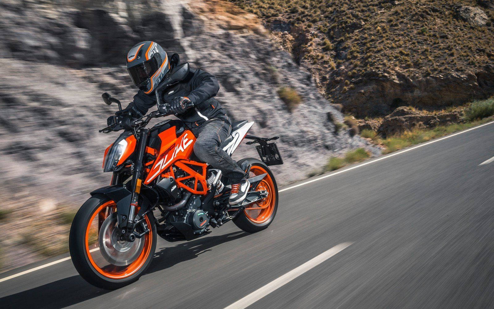 機車 桌布 壁紙 — 一名身穿黑色裝備的騎士，在鮮橙色KTM Duke摩托車上傾斜入彎，沿著蜿蜒的山路飛馳。