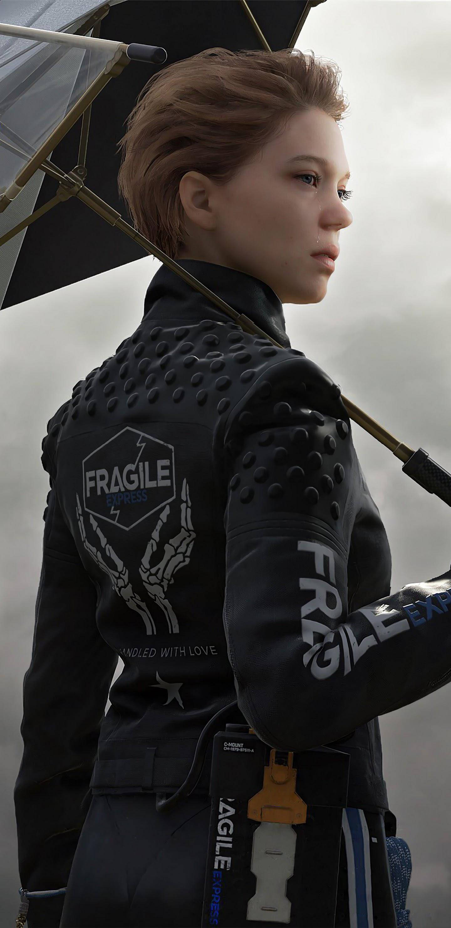 蕾雅·瑟杜 桌布 壁紙 — 一名身穿黑色裝甲外套、帶有'FRAGILE'字樣的女性，在灰色天空下撐著雨傘。