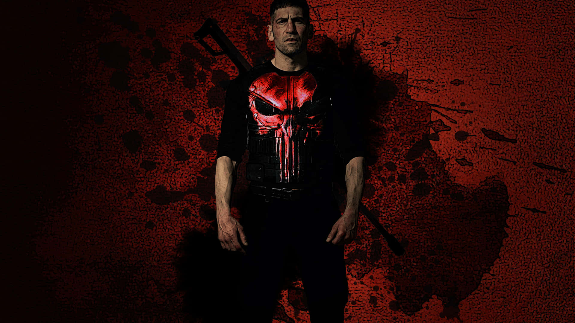 Punisher Desktop 桌布 壁紙 — 一名身穿黑色襯衫、帶有紅色骷髏標誌的男子，站在帶有深色飛濺紋理的紅色背景前。