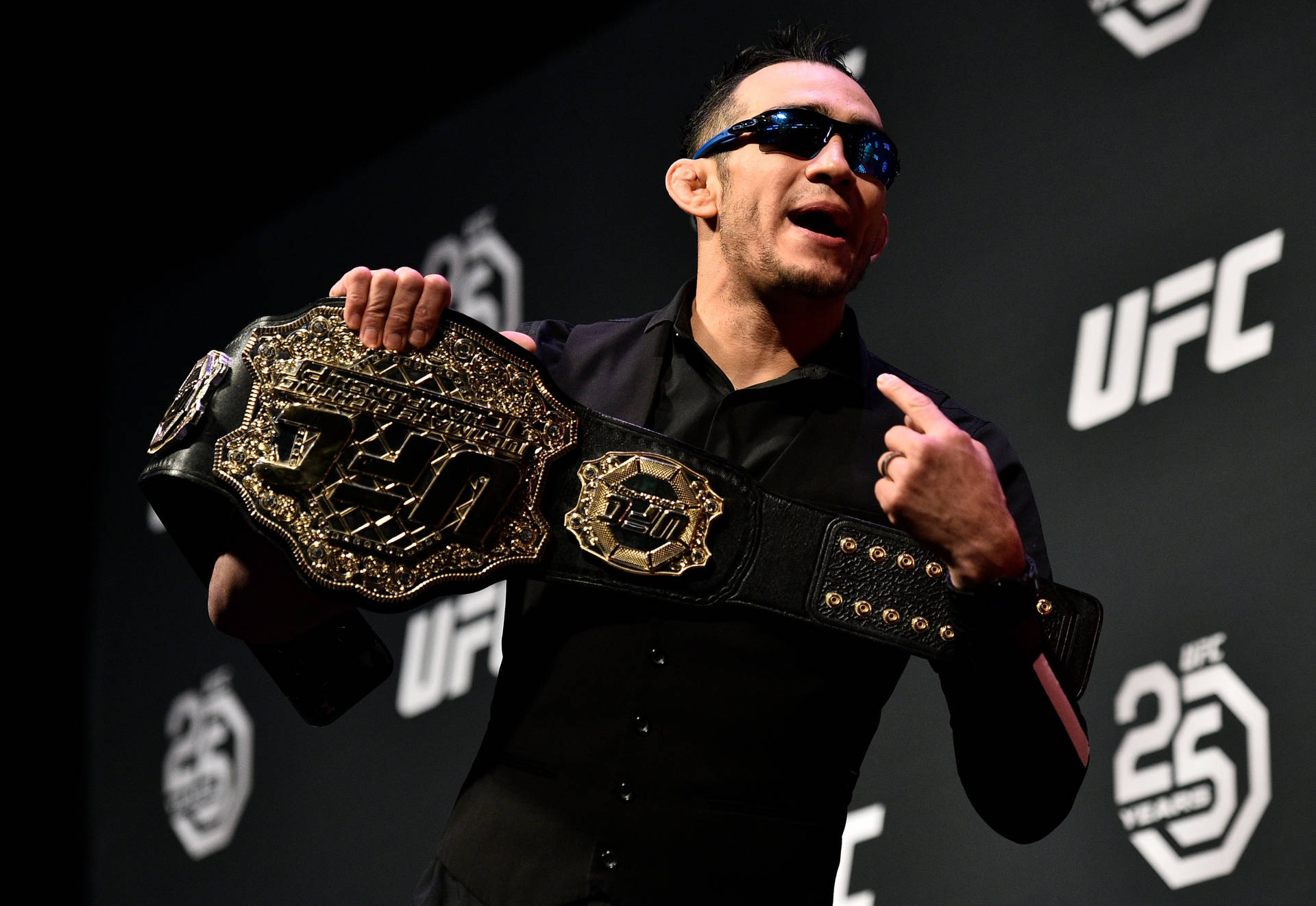 UFC 桌布 壁紙 — 一名身穿黑色襯衫和太陽眼鏡的戰士，在深色品牌背景前自豪地展示一條金色的UFC冠軍腰帶。