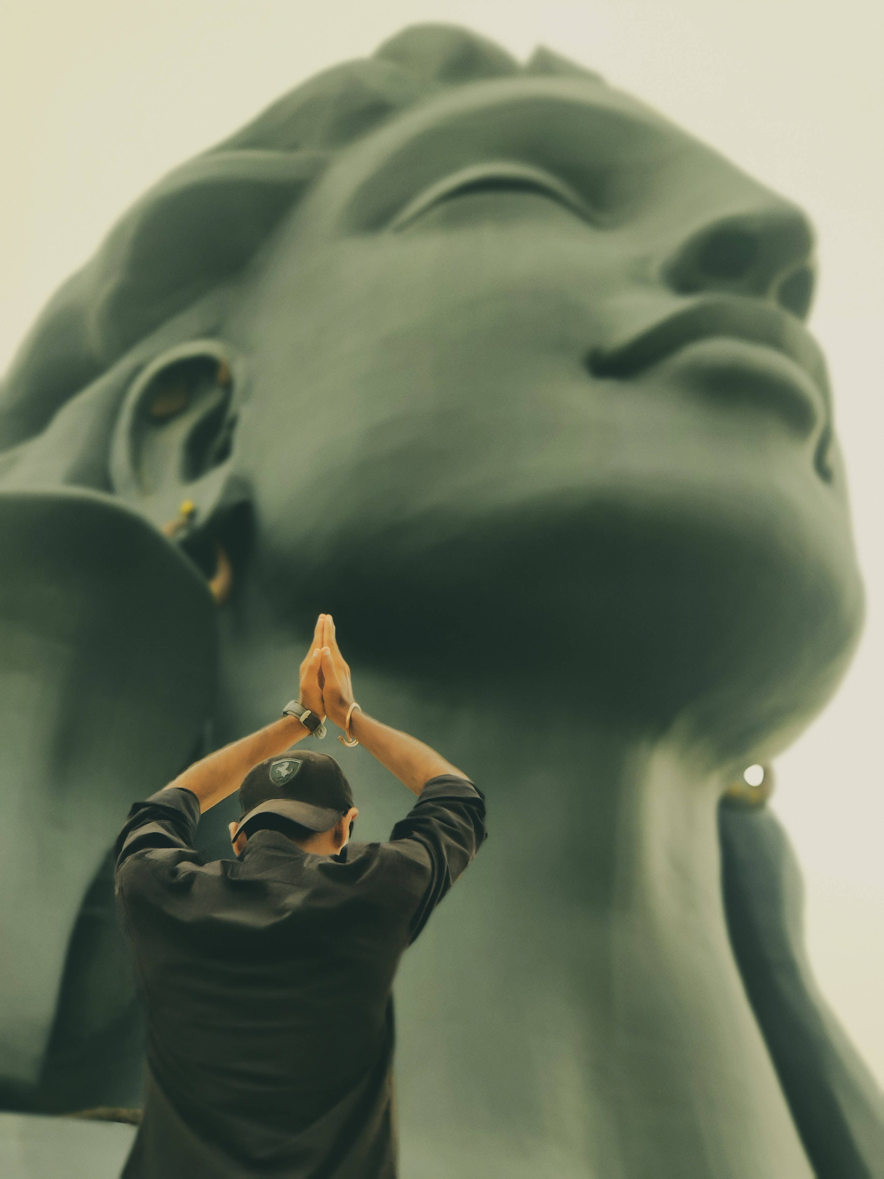 adiyogi shiva 桌布 壁紙 — 一名身穿黑色襯衫和帽子的人，在柔和漫射光線下，雙手合十，在一尊巨大而寧靜的佛像前祈禱。