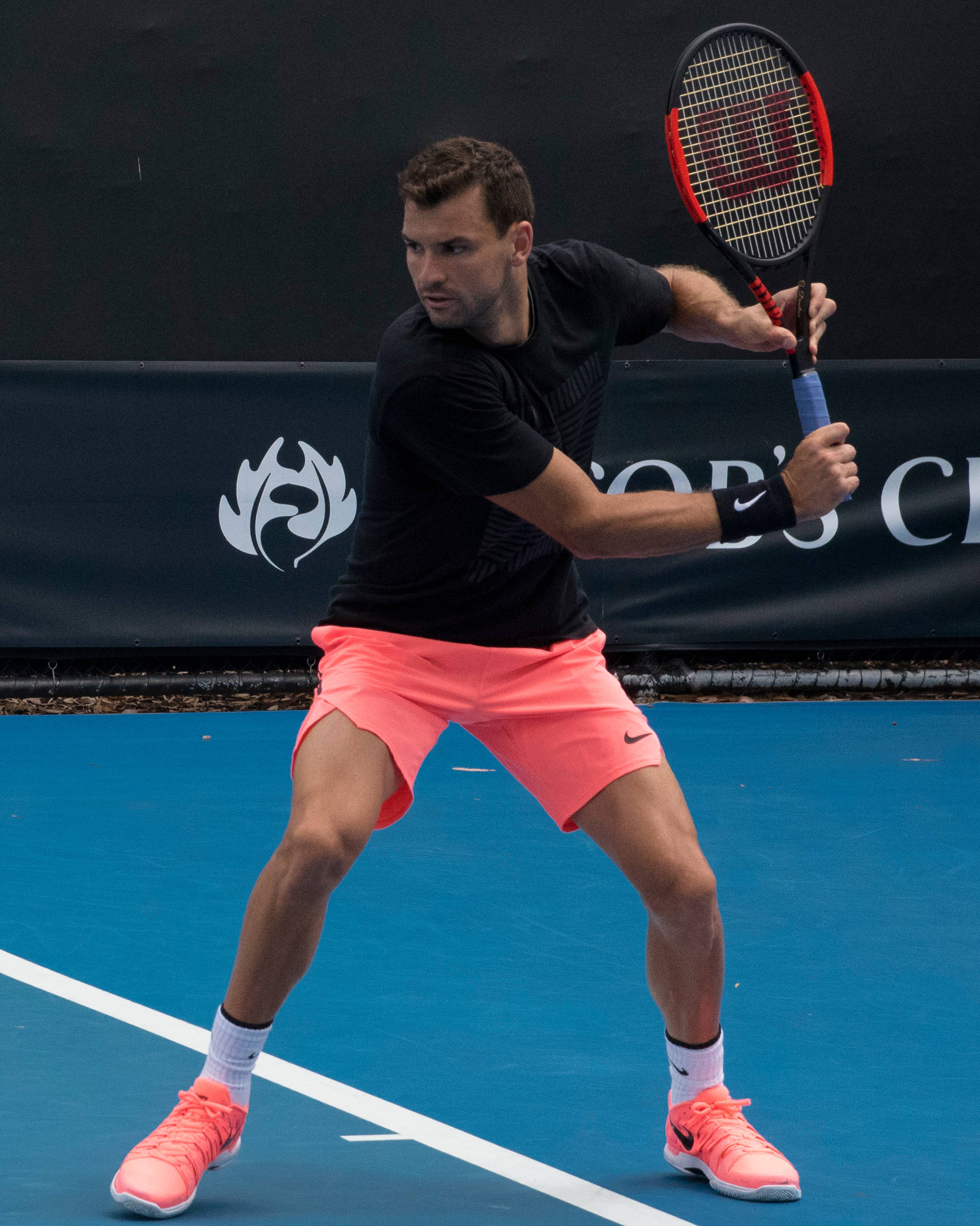 Tennis 桌布 壁紙 — 一名身穿黑色襯衫和珊瑚色短褲的男性網球運動員在藍色硬地球場上專注地揮拍。