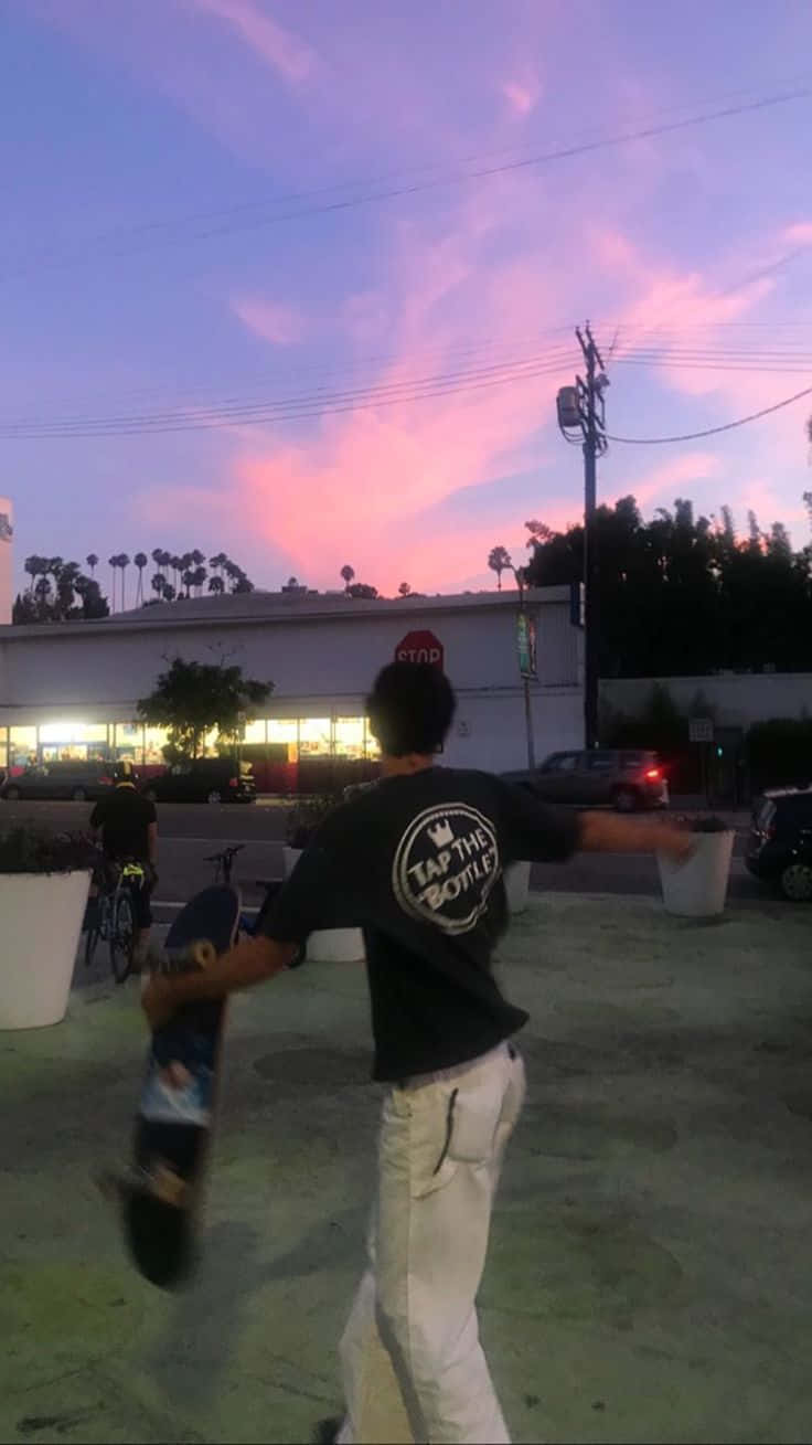 skateboarding 桌布 壁紙 — 一名身穿黑色襯衫和白色褲子的滑板者在黃昏時分在混凝土廣場上保持平衡，背景是粉紫色天空和棕櫚樹。