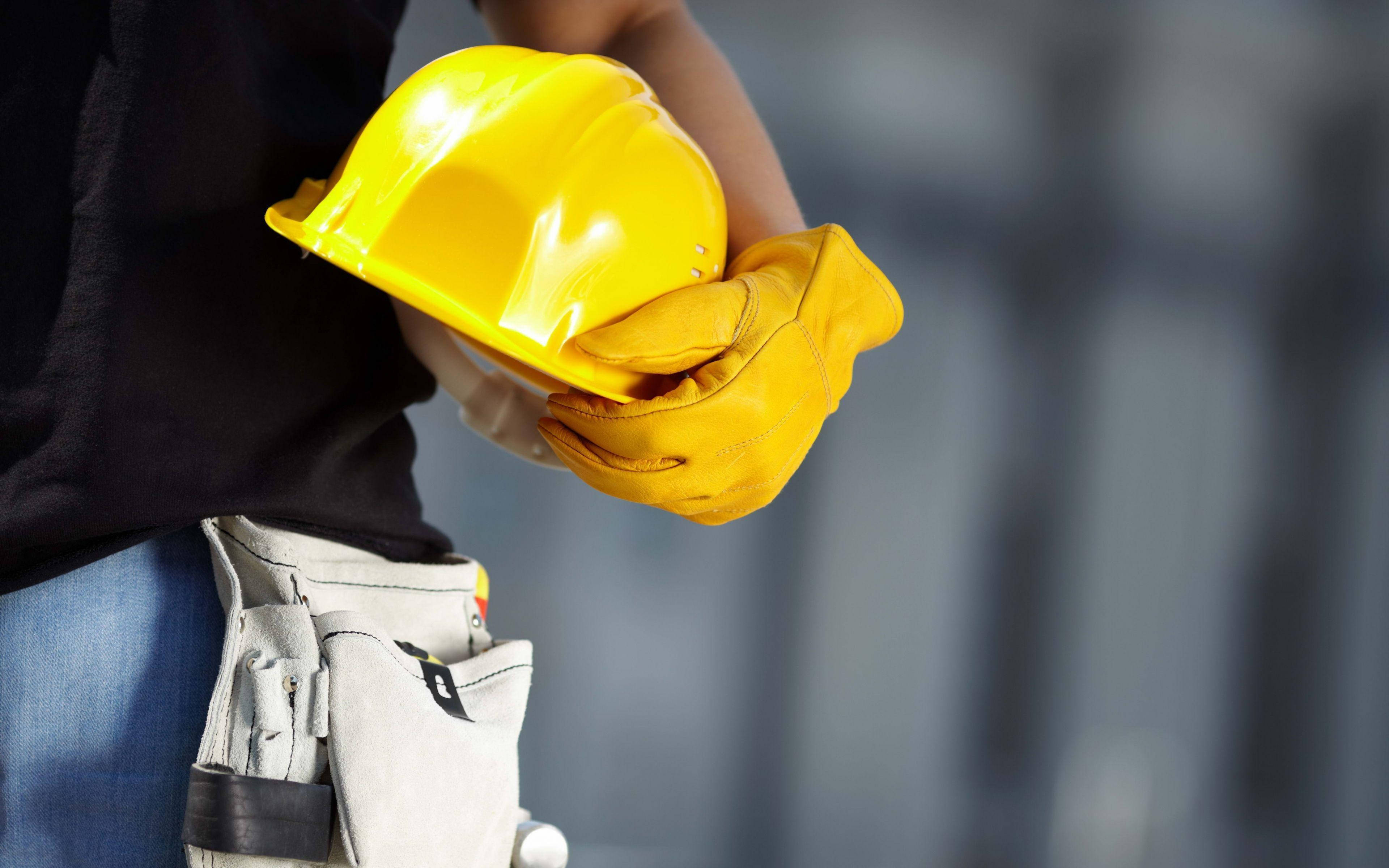 construction 桌布 壁紙 — 一名身穿黑色襯衫和藍色牛仔褲的工人，手持亮黃色安全帽和黃色手套，並佩戴工具腰帶。