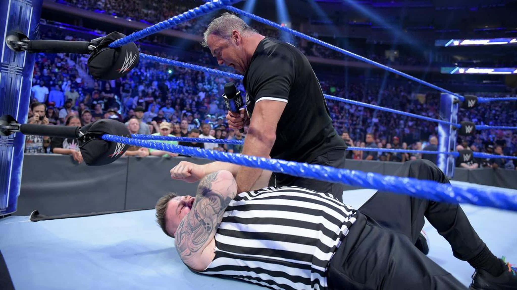 WWE 桌布 壁紙 — 一名身穿黑色襯衫的摔角手攻擊一名身穿條紋襯衫、躺在藍色摔角墊上的裁判，場地燈光明亮。
