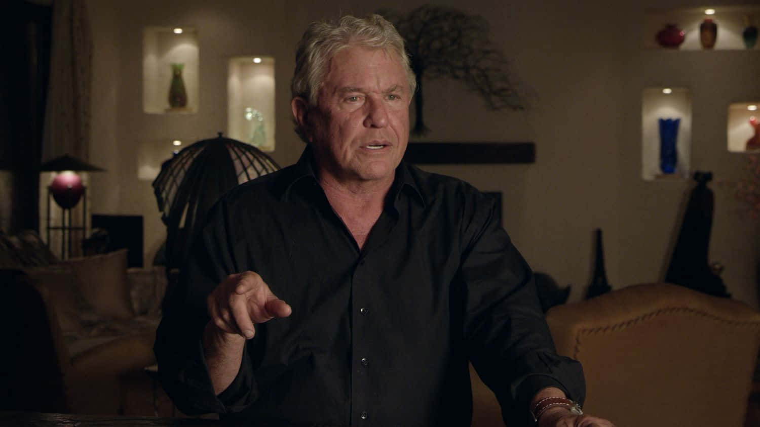 Tom Berenger 桌布 壁紙 — 一名身穿黑色襯衫的男子在光線昏暗的現代客廳中說話時做手勢，客廳設有環境照明和裝飾架。