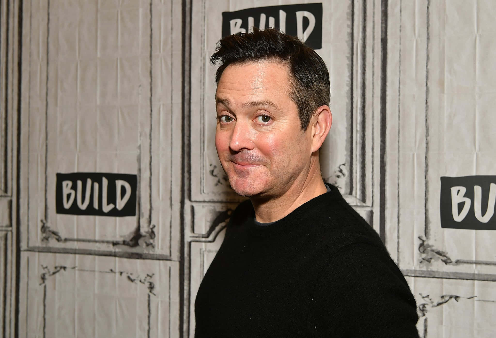 thomas lennon 桌布 壁紙 — 一名身穿黑色襯衫的男子在帶有 'BUILD' 標誌的紋理牆前頑皮地微笑。
