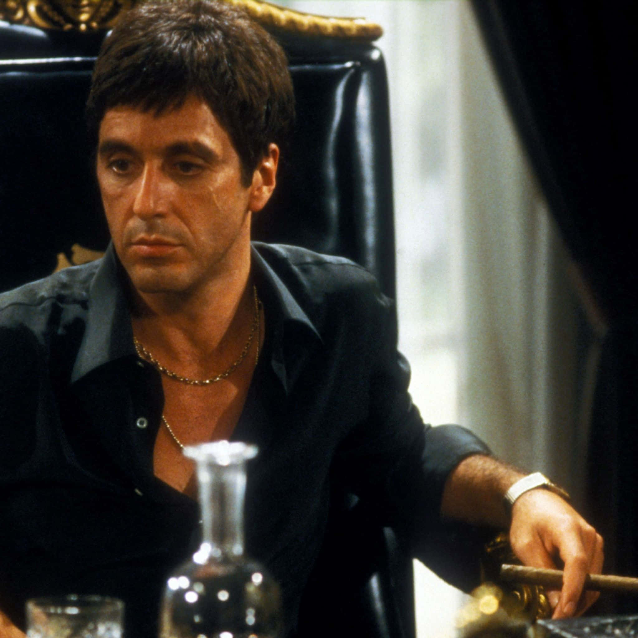 tony montana 桌布 壁紙 — 一名身穿黑色襯衫的男子坐在皮椅上，手持雪茄，表情嚴肅。