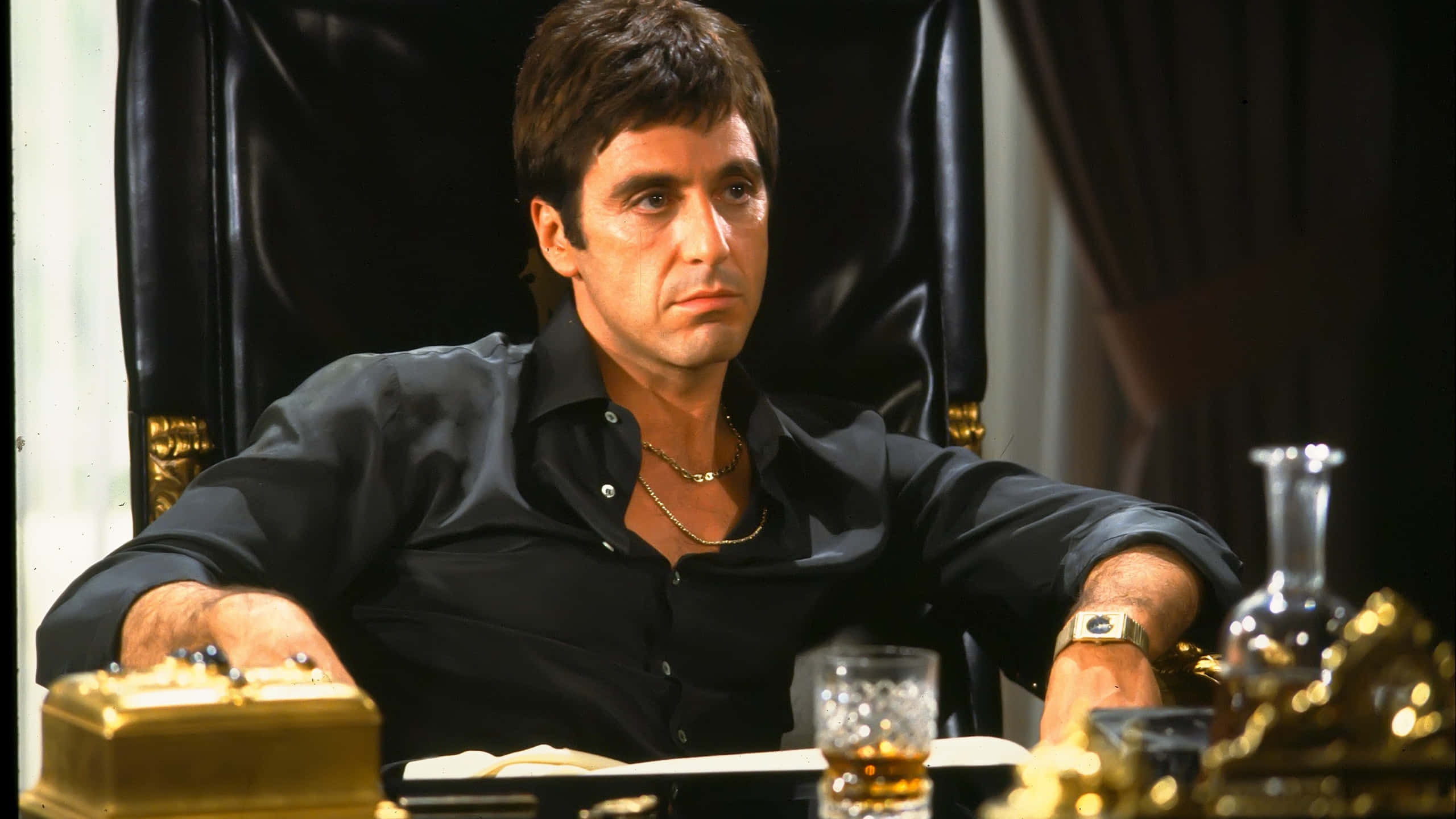 tony montana 桌布 壁紙 — 一名身穿黑色襯衫的男子威嚴地坐在皮椅上，周圍環繞著金色物品和一杯威士忌。