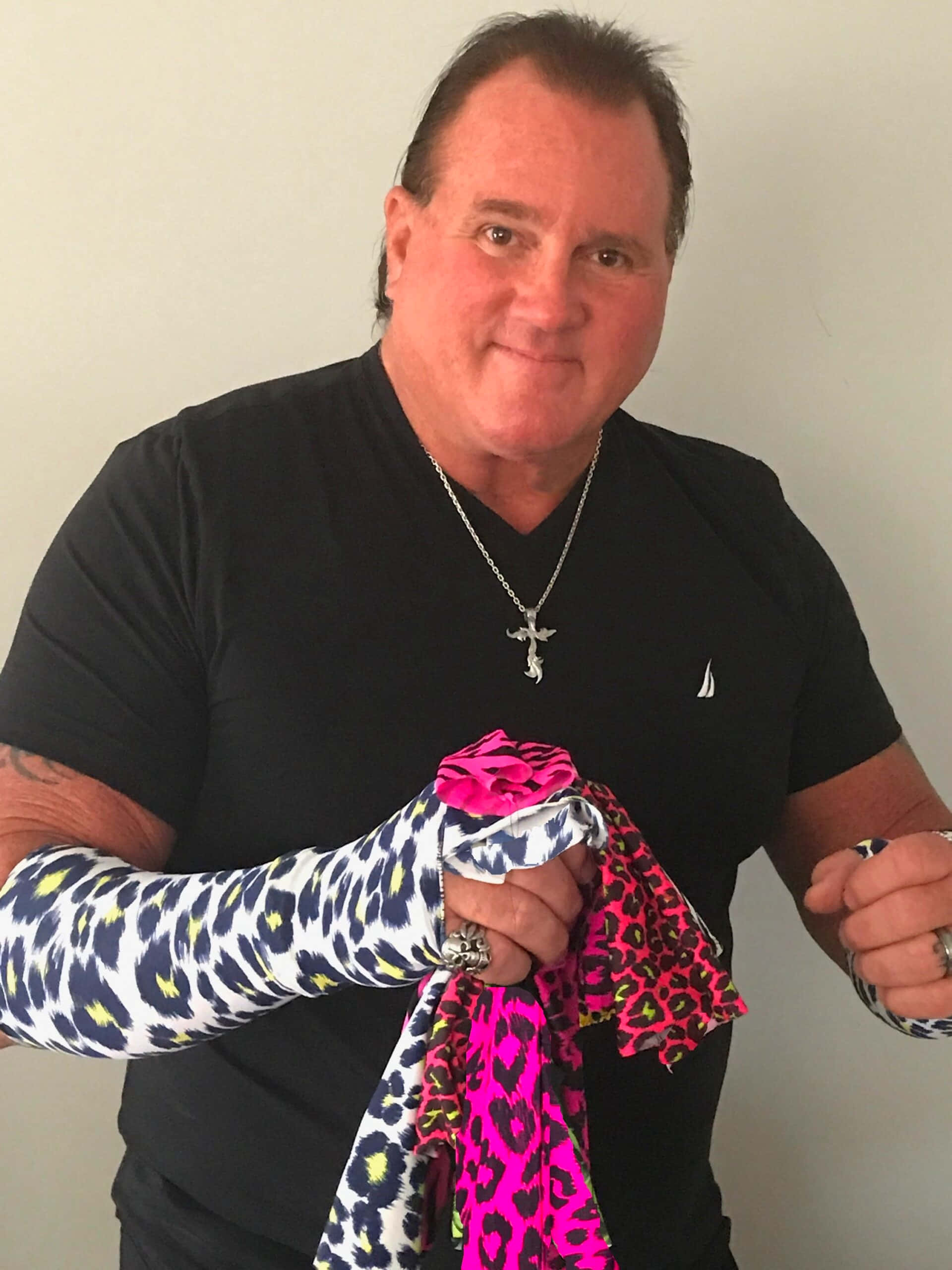 Brutus Beefcake 桌布 壁紙 — 一名身穿黑色襯衫的男子微笑著，手持鮮豔的粉紅色和藍色豹紋布料靠在素牆上。