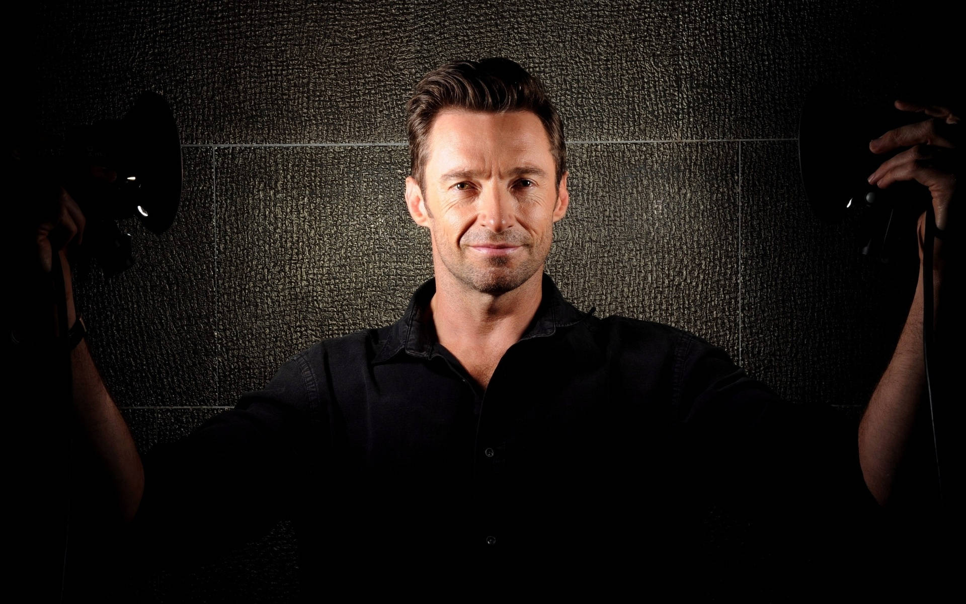 Hugh Jackman 桌布 壁紙 — 一名身穿黑色襯衫的男子站在中央，背景是深色紋理牆壁，兩側的攝影棚燈光將他框住。