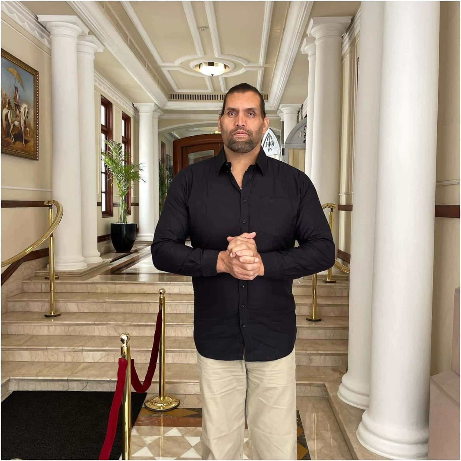Indian 桌布 壁紙 — 一名身穿黑色襯衫的男子自信地站在宏偉的大理石樓梯頂端，兩側是白色圓柱和紅色天鵝絨繩索。