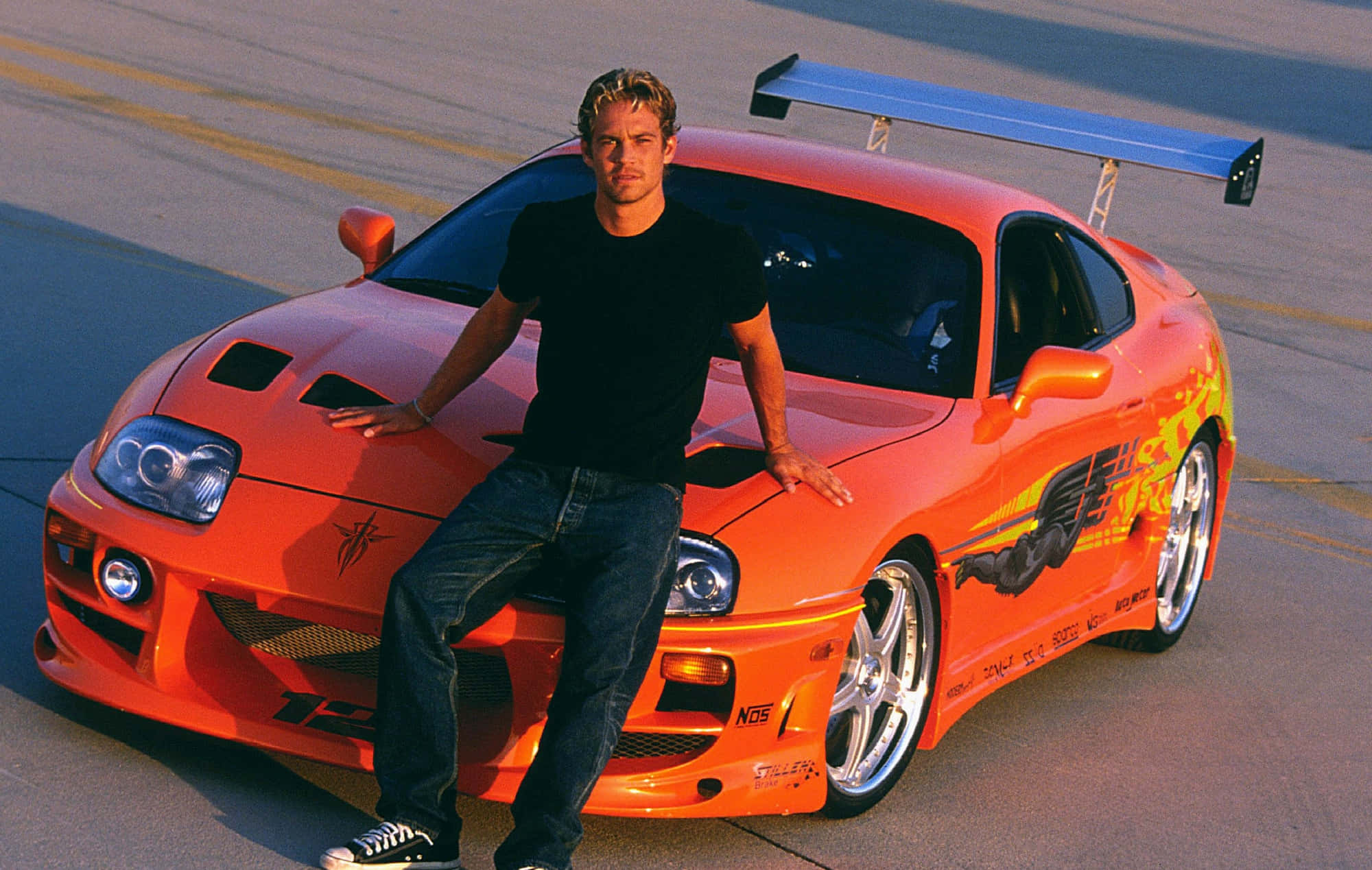 Fast And Furious 桌布 壁紙 — 一名身穿黑色襯衫的男子自信地靠在一輛亮橙色改裝跑車上，車身有藍色尾翼和火焰貼紙，停在鋪砌的停車場上。