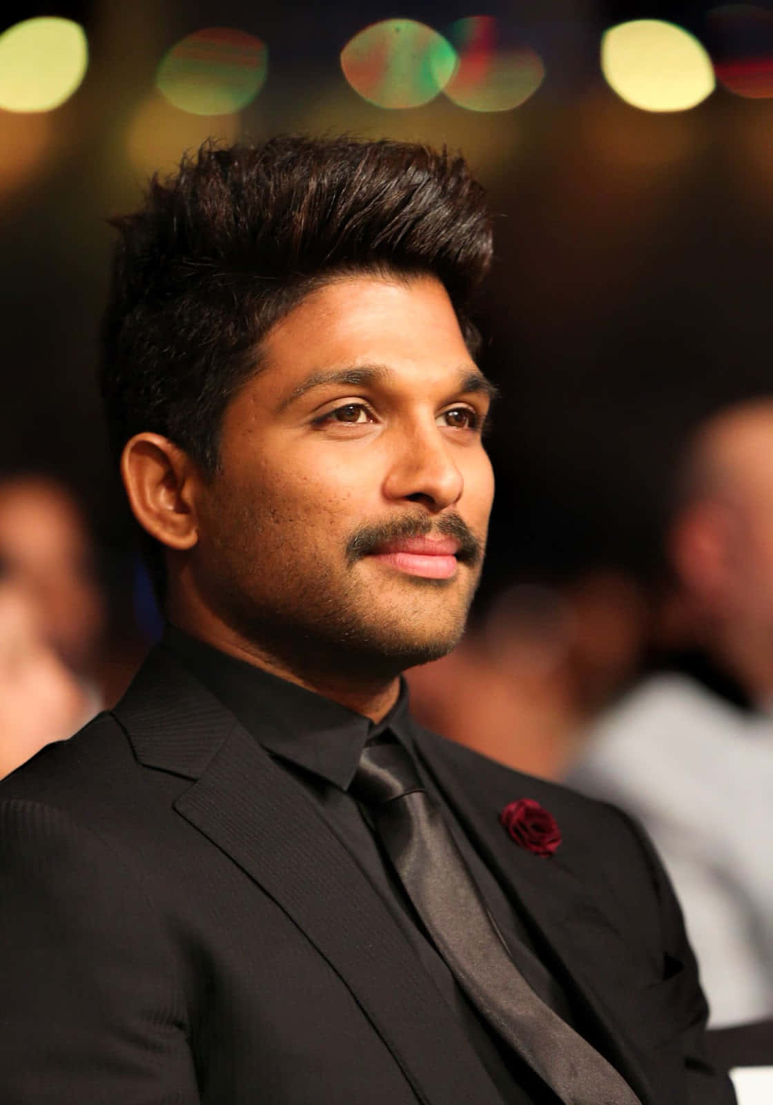 Allu Arjun 桌布 壁紙 — 一名身穿黑色西裝、佩戴紅色玫瑰胸針的男子，在模糊的散景背景前沉思凝視。