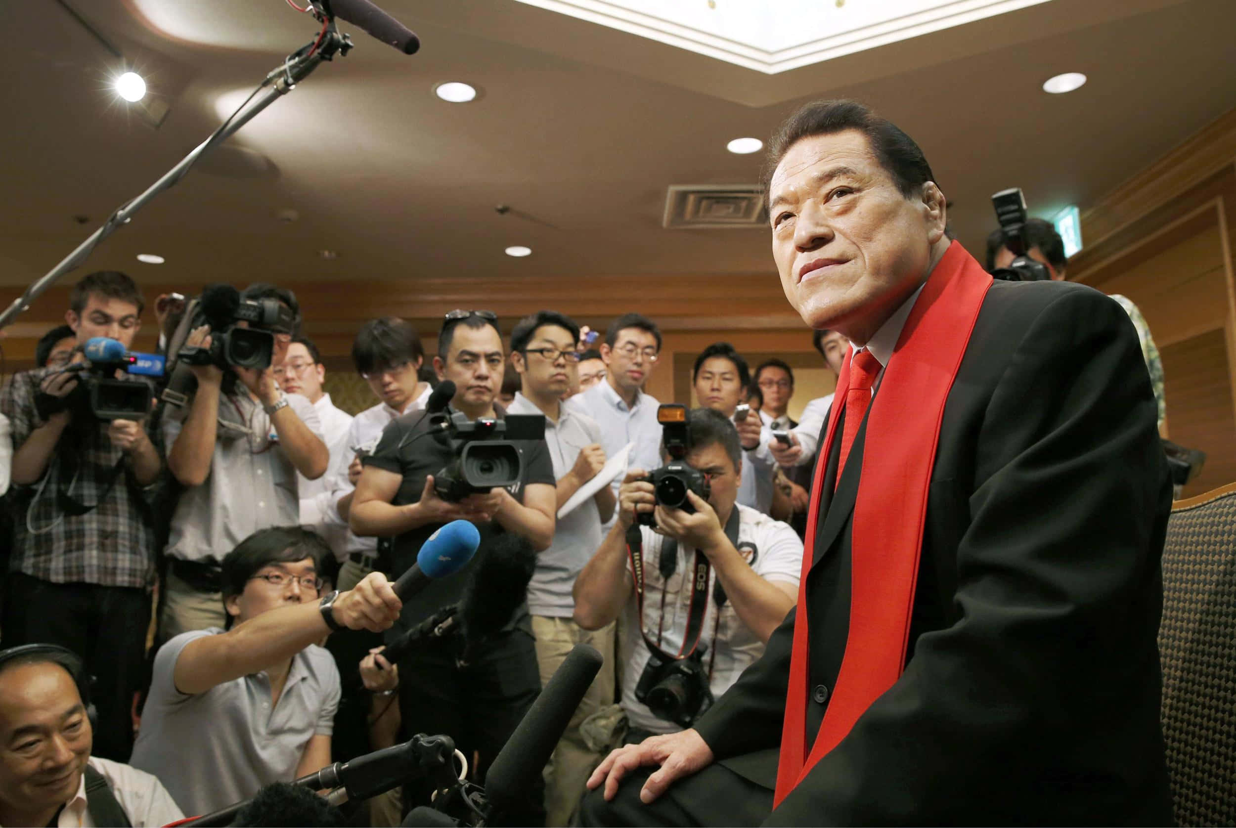 運動 桌布 壁紙 — 一名身穿黑色西裝、戴著大膽紅色圍巾的男子在記者室內平靜地坐著，周圍是記者和攝影機。
