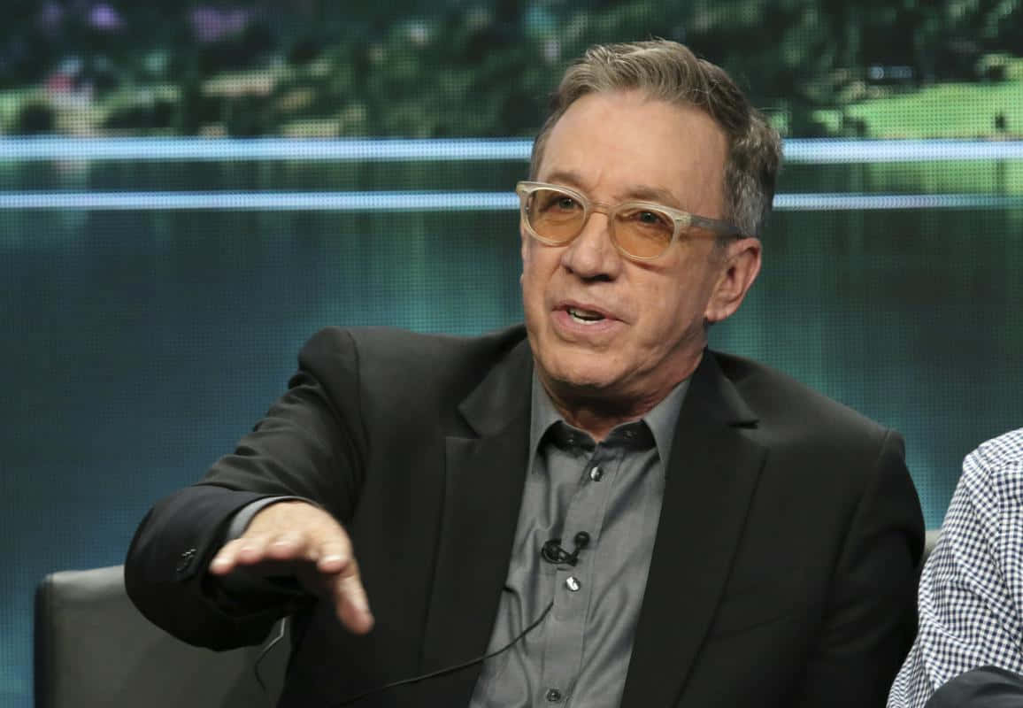 Tim Allen 桌布 壁紙 — 一名身穿黑色西裝、戴黃色眼鏡的男子在模糊綠色螢幕背景的座談會上發言時做手勢。
