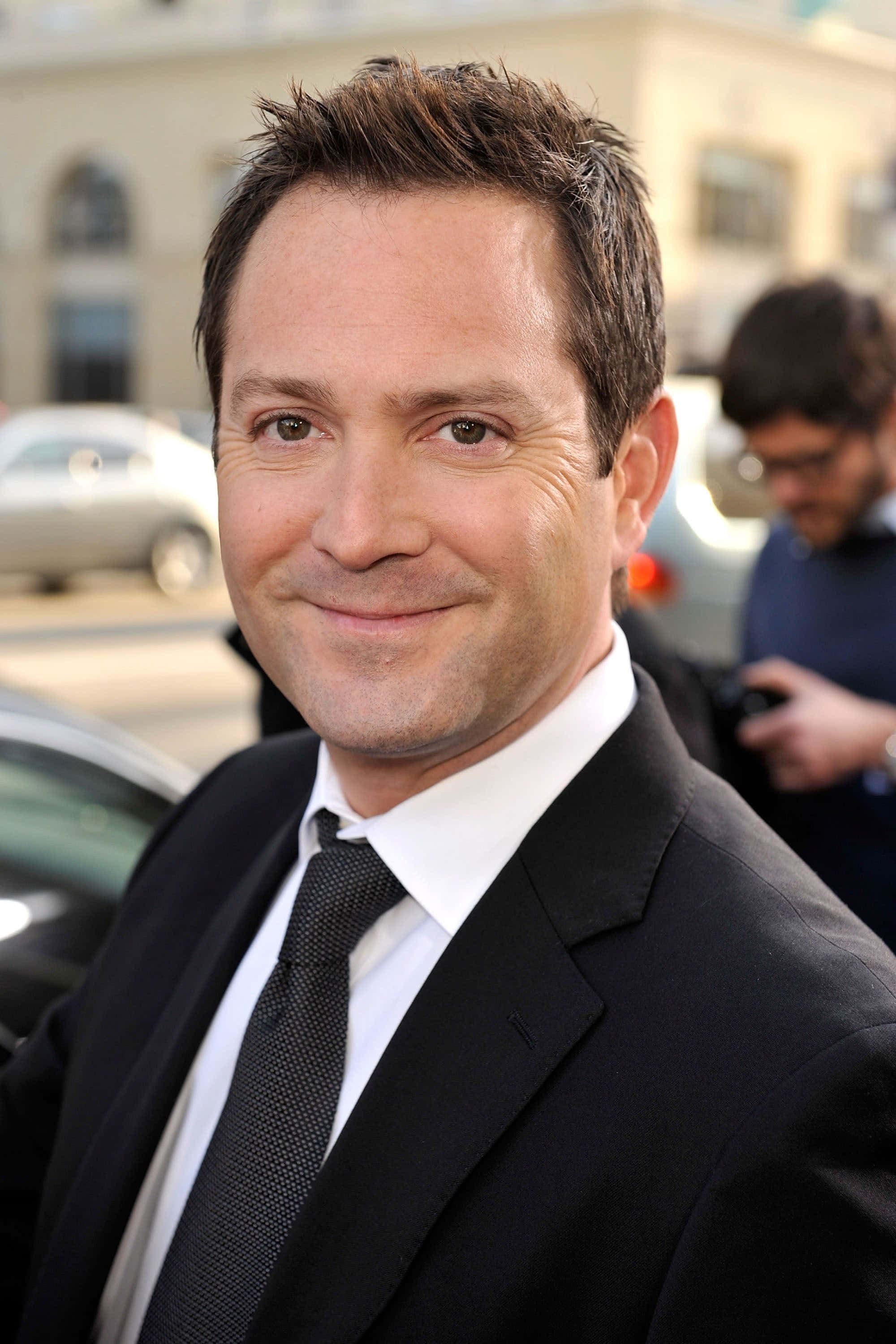 thomas lennon 桌布 壁紙 — 一名身穿黑色西裝及領帶的男子，在城市街道上對著鏡頭溫暖微笑，背景是汽車和建築物，呈現柔和焦點。