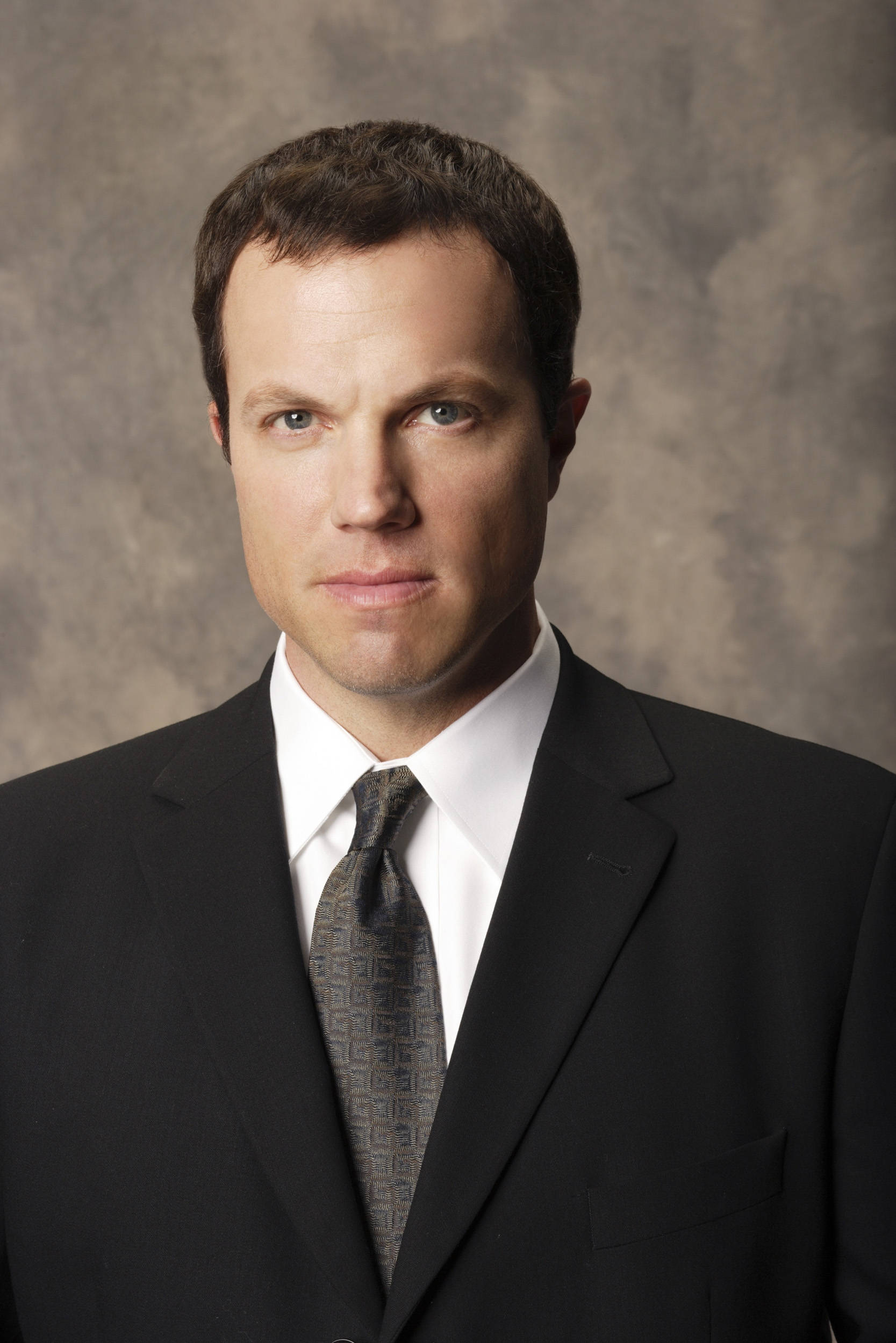 adam baldwin 桌布 壁紙 — 一名身穿黑色西裝和圖案領帶的男子，在紋理灰色背景前直視鏡頭。
