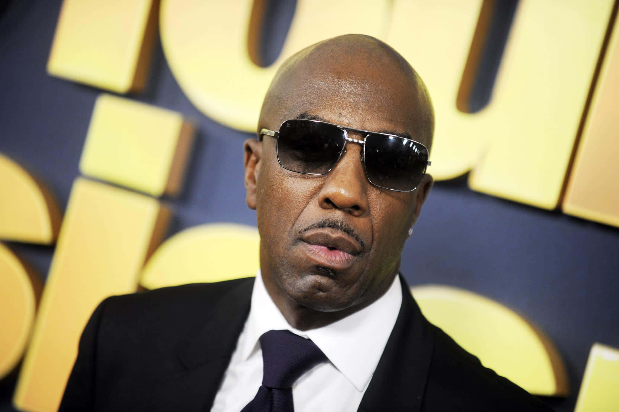 J.b. Smoove 桌布 壁紙 — 一名身穿黑色西裝和太陽眼鏡的男子在鮮明的黃色和藍色背景前自信地擺姿勢。