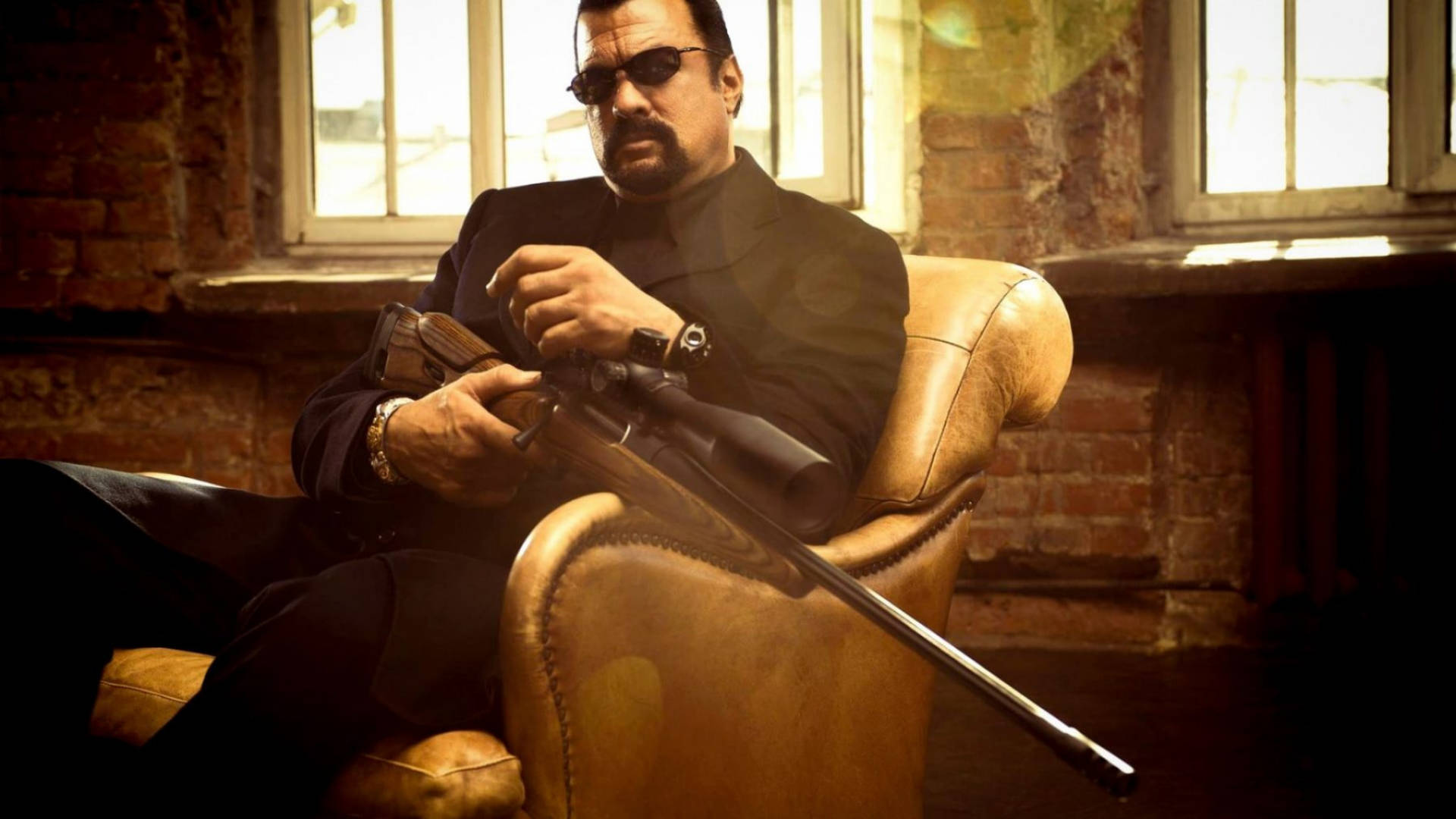 Steven Seagal 桌布 壁紙 — 一名身穿黑色西裝和太陽眼鏡的男子坐在皮扶手椅上，在陽光照射的磚房裡手持狙擊步槍。