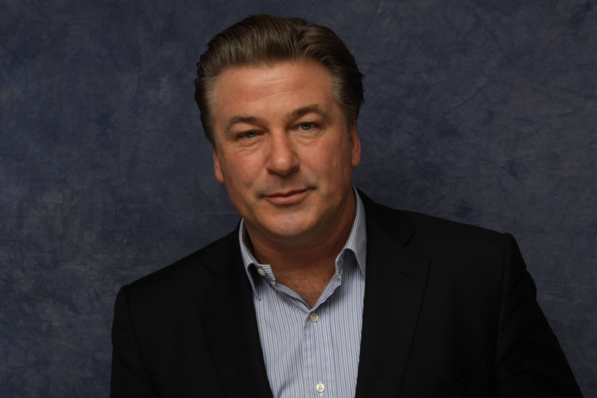 Alec Baldwin 桌布 壁紙 — 一名身穿黑色西裝和條紋襯衫的男子，在帶有紋理的藍色背景前擺出冷靜的表情。