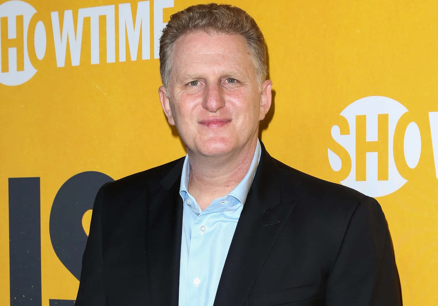 Michael Rapaport 桌布 壁紙 — 一名身穿黑色西裝和淺藍色襯衫的男子，在明亮的黃色Howtime背景前對著鏡頭微笑。
