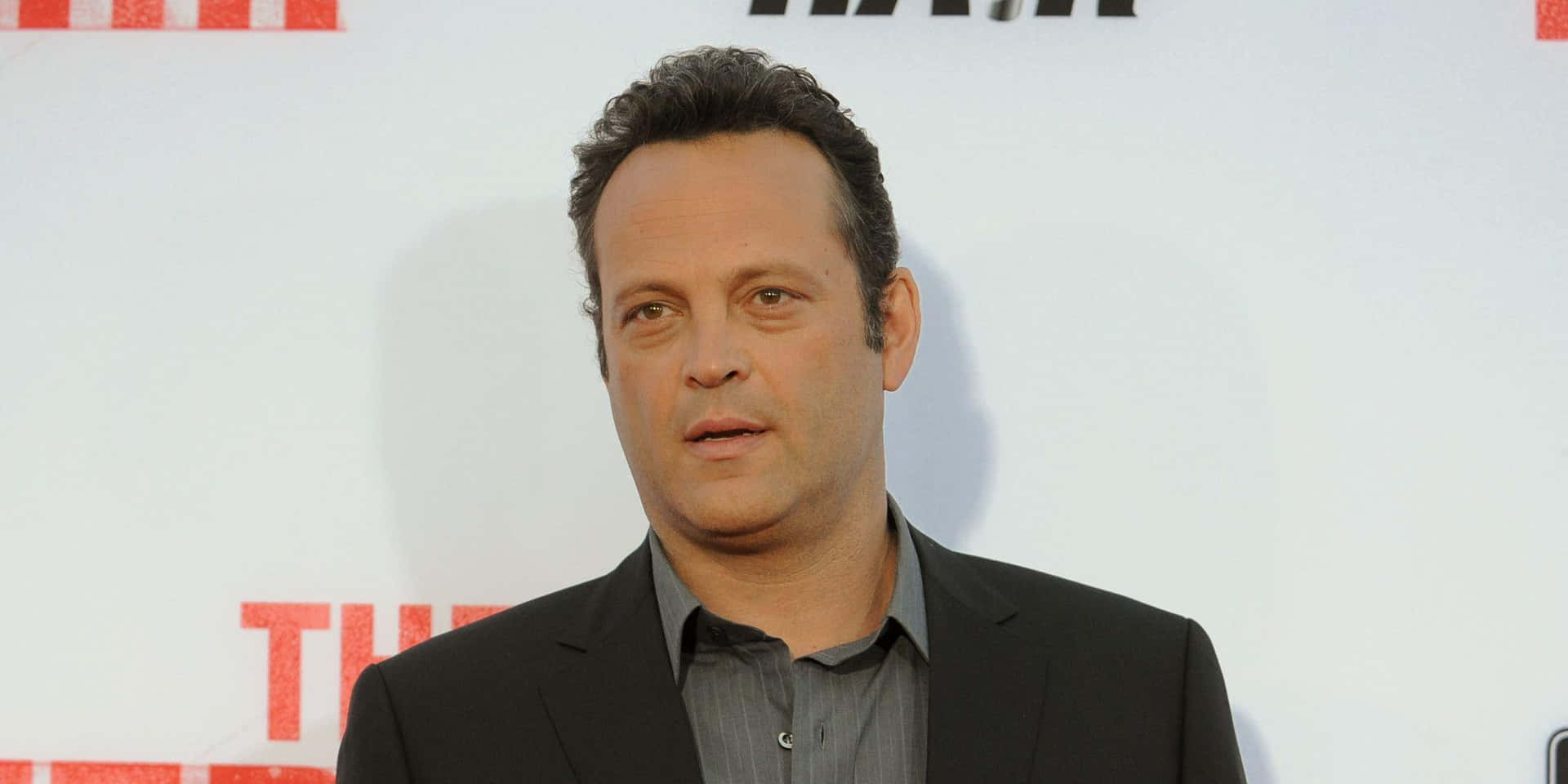 Vince Vaughn 桌布 壁紙 — 一名身穿黑色西裝和灰色襯衫的男子站在白色背景前，背景上有紅色字樣。