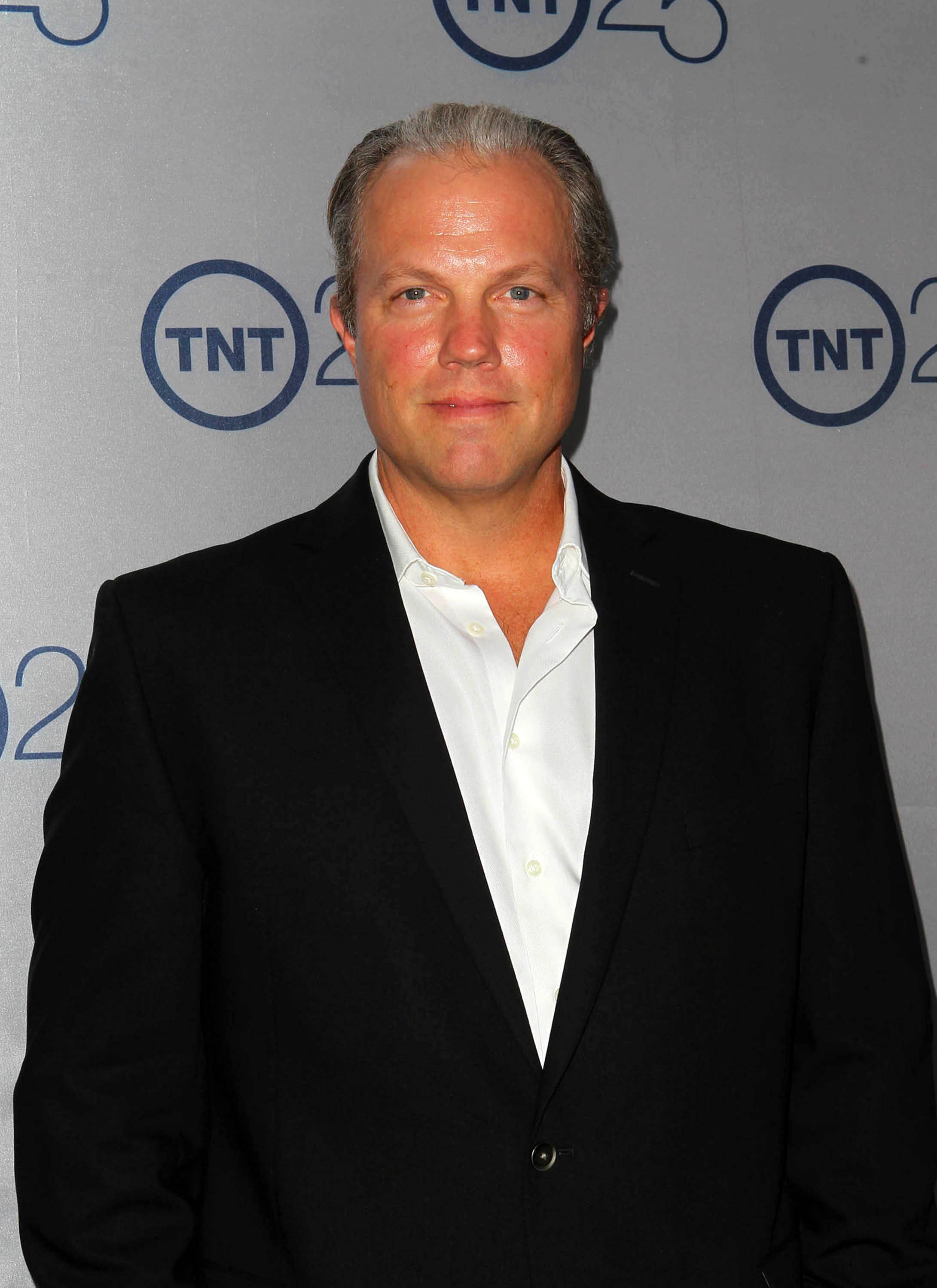 adam baldwin 桌布 壁紙 — 一名身穿黑色西裝和白色襯衫的男子自信地站在灰色TNT 25週年背景前。