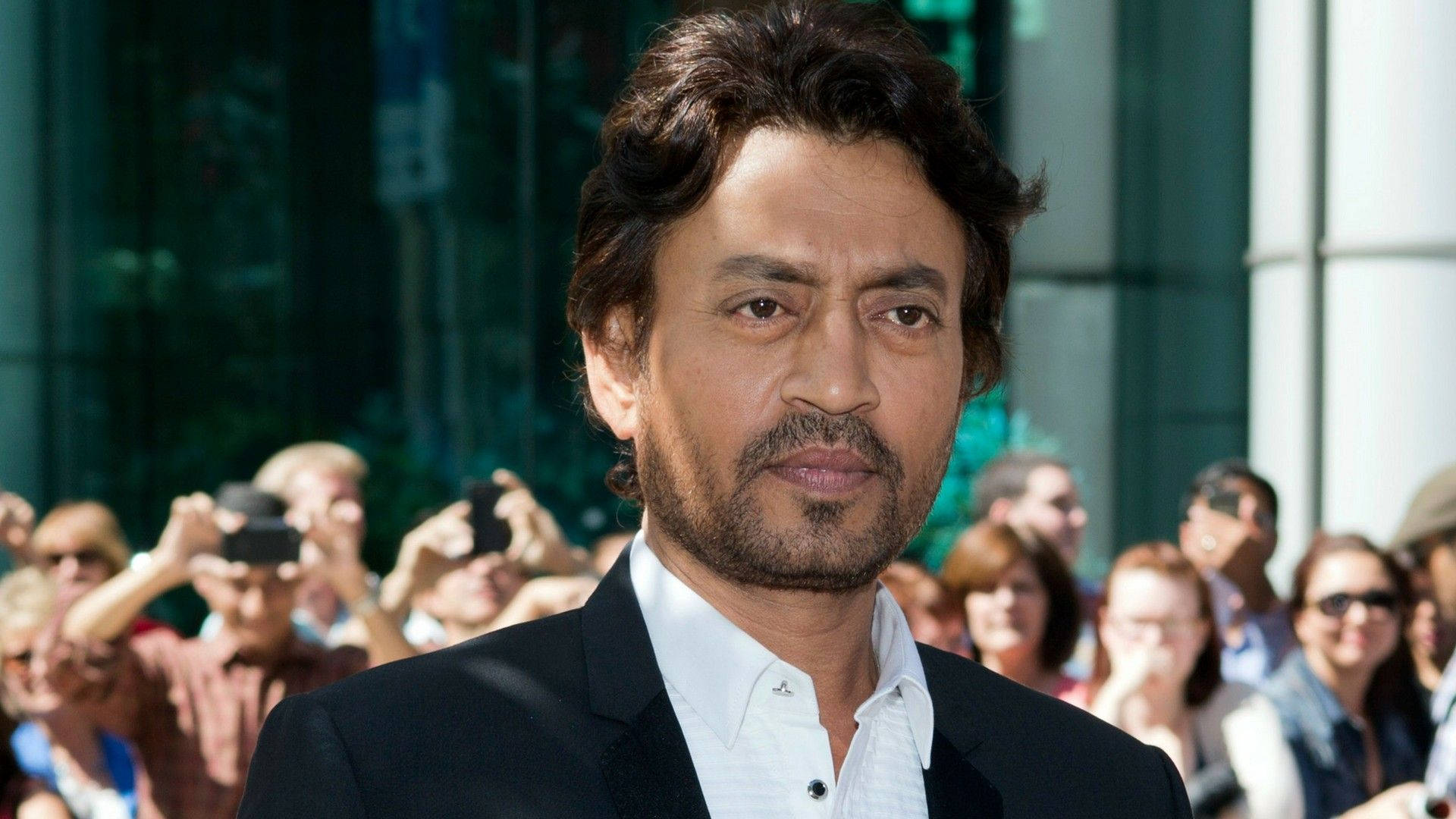 Irrfan Khan 桌布 壁紙 — 一名身穿黑色西裝和白色襯衫的男子自信地站在玻璃建築外的一群攝影師面前。