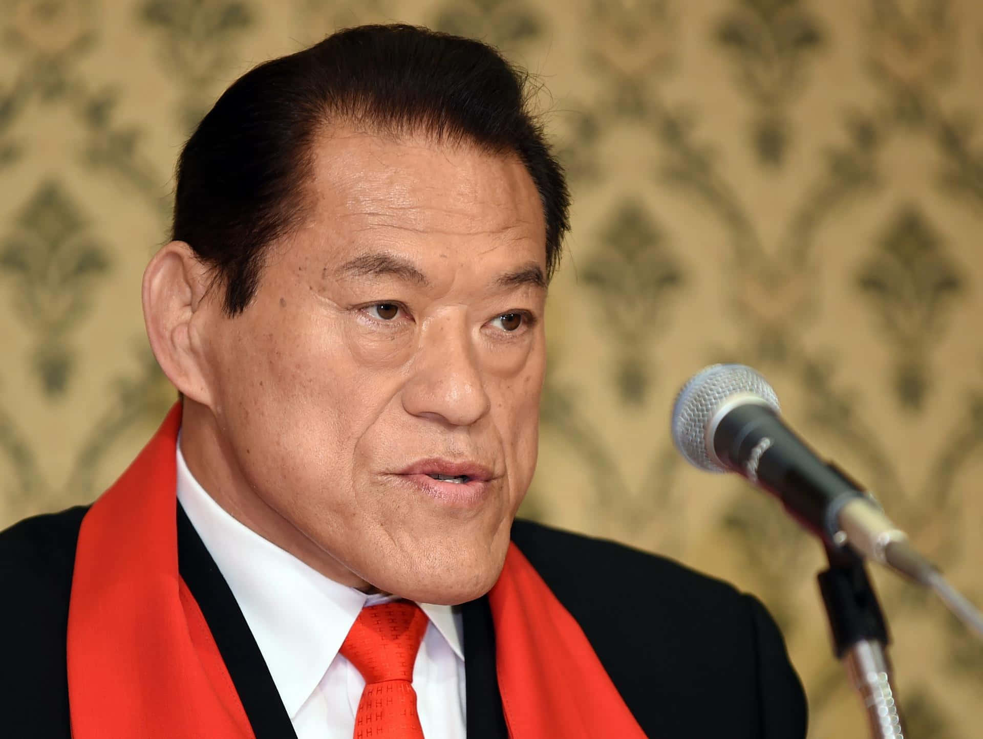 運動 桌布 壁紙 — 一名身穿黑色西裝和紅色領帶的男子在圖案壁紙背景前對著麥克風說話的特寫。