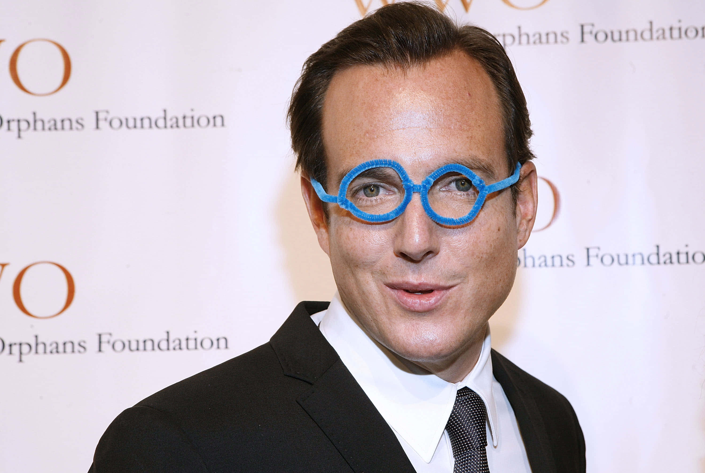 Will Arnett 桌布 壁紙 — 一名身穿黑色西裝和領帶的男子在慈善活動中戴著超大的亮藍色新奇眼鏡微笑。
