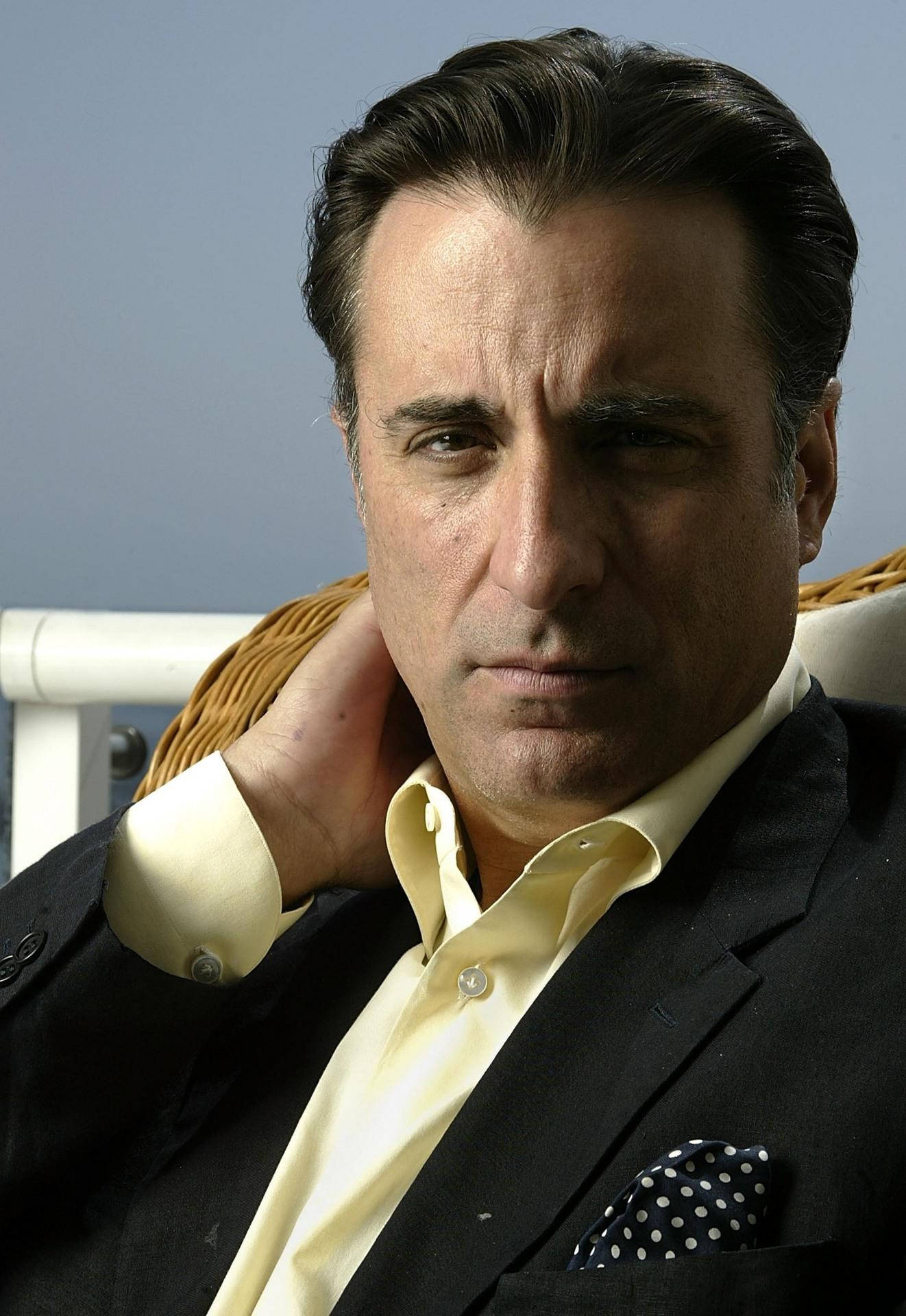 Andy Garcia 桌布 壁紙 — 一名身穿黑色西裝和黃色襯衫的男子，表情嚴肅地將頭靠在手上，倚著藤椅。