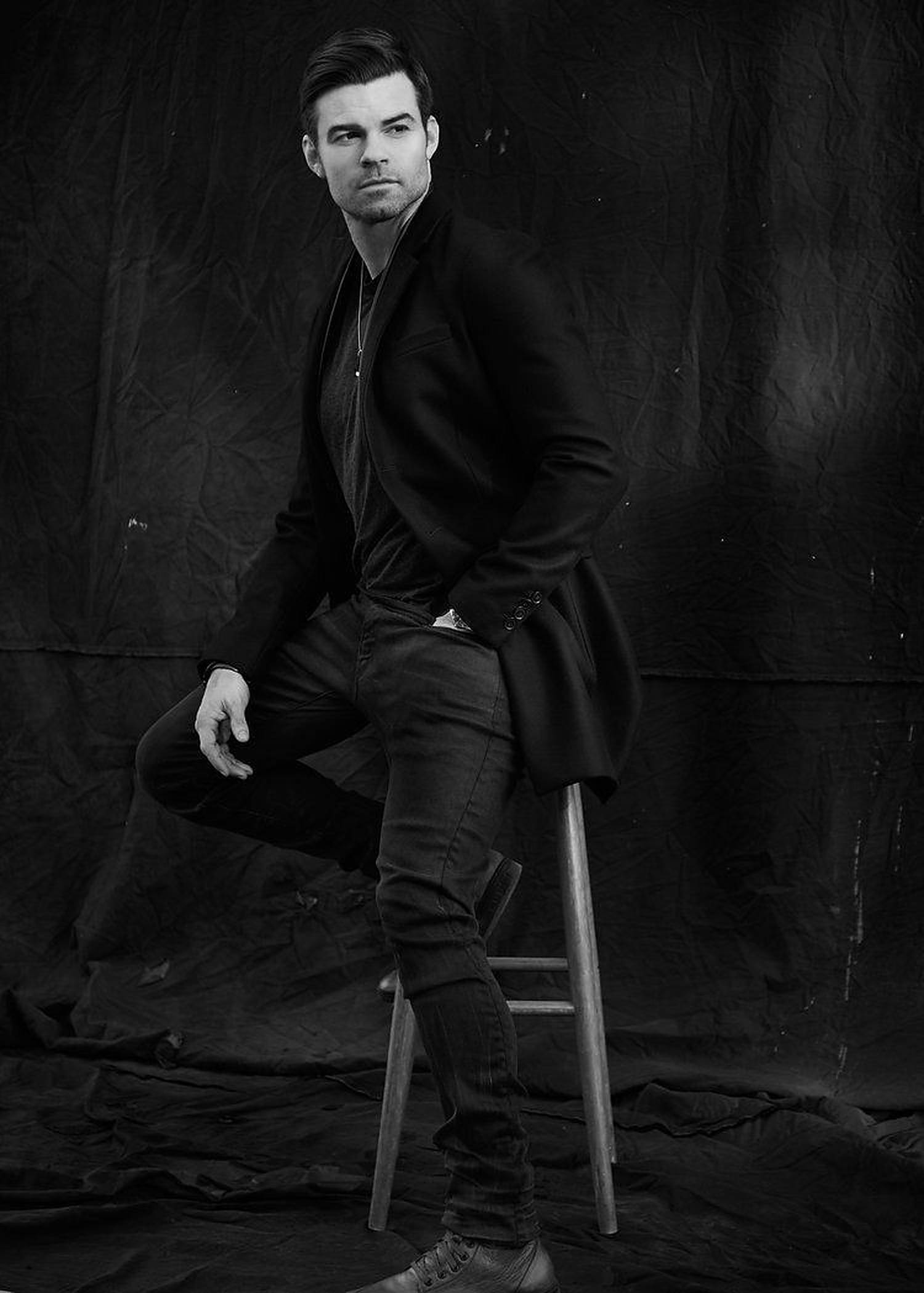 Daniel Gillies 桌布 壁紙 — 一名身穿黑色西裝外套和牛仔褲的男子，自信地坐在木凳上，背景是帶有紋理的黑色，營造出陰鬱的單色工作室肖像。