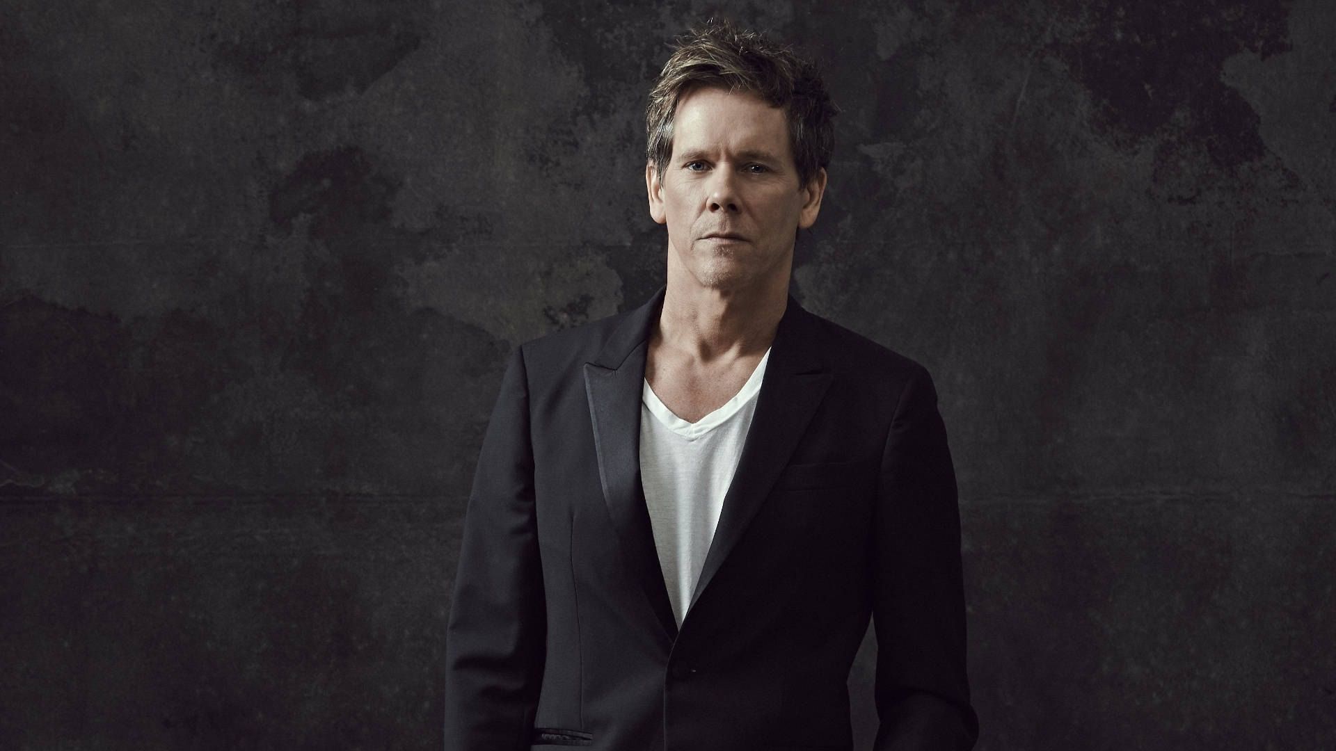 actor 桌布 壁紙 — 一名身穿黑色西裝外套和白色T恤的男子，靠著質感粗糙的深灰色牆壁，表情嚴肅。