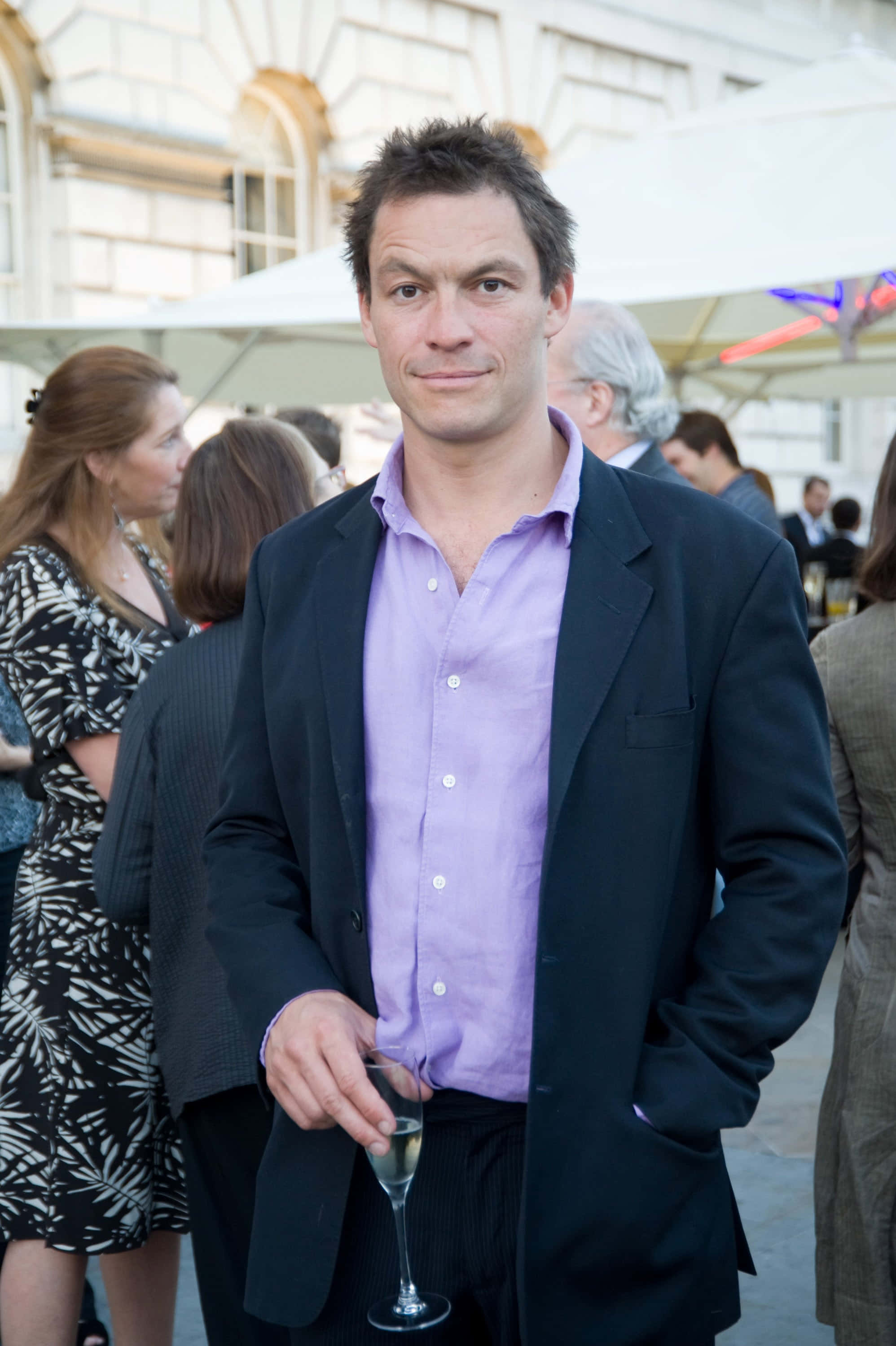Dominic West 桌布 壁紙 — 一名身穿黑色西裝外套和紫色襯衫的男子，在白色遮陽傘下的戶外社交聚會中手持香檳杯。