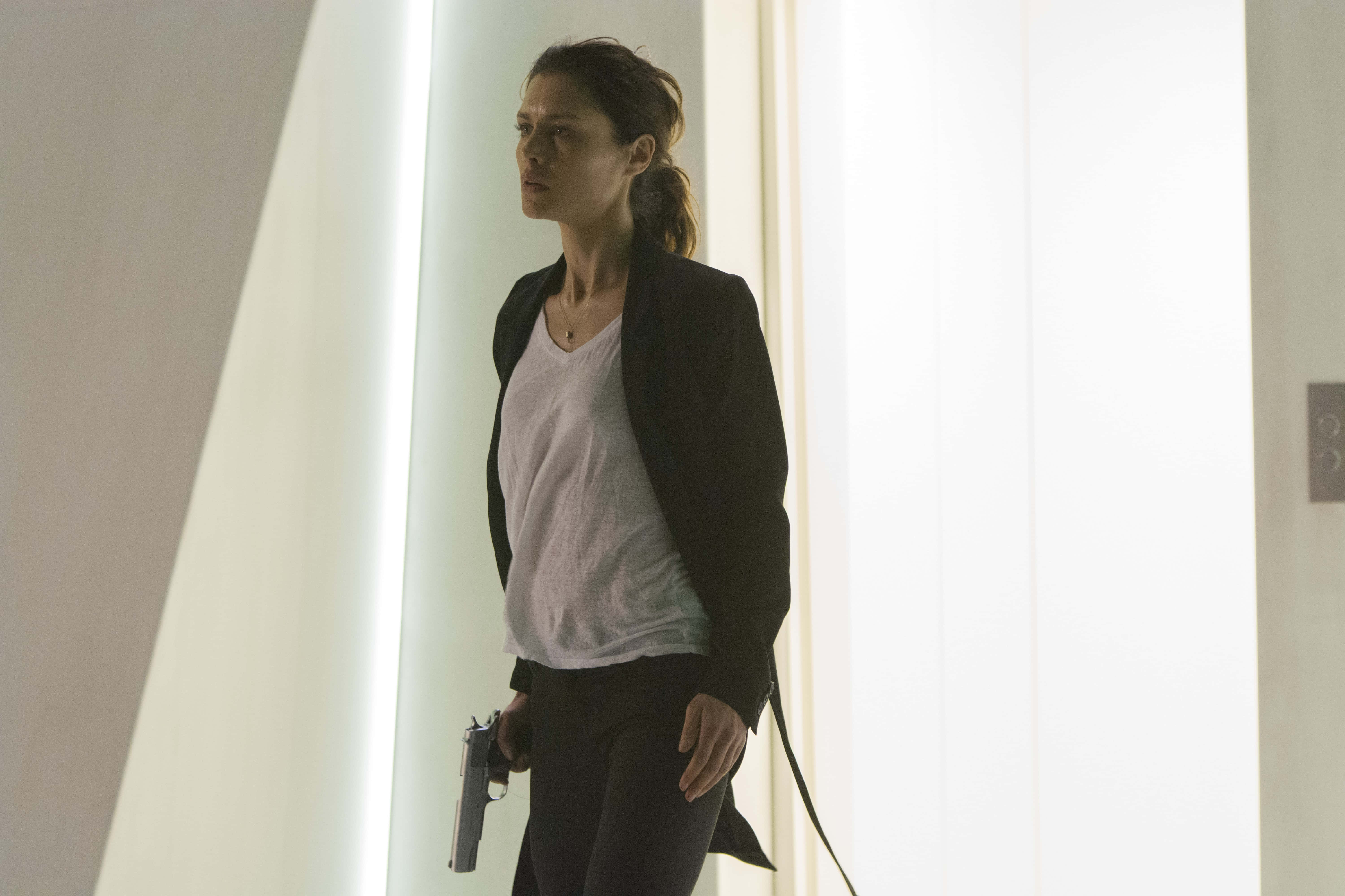 Hitman Agent 47 桌布 壁紙 — 一名身穿黑色西裝外套的女子站在光線明亮的現代走廊中，手持手槍。