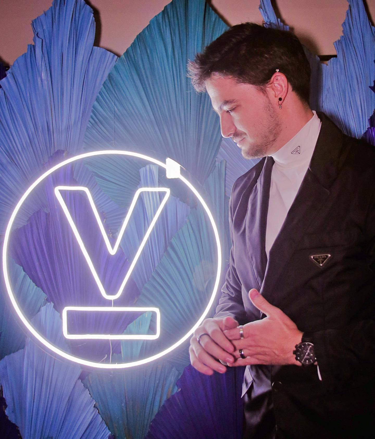 Man In Suit 桌布 壁紙 — 一名身穿黑色西裝外套的男子站在發光的霓虹V標誌旁邊，背景是帶紋理的藍色。