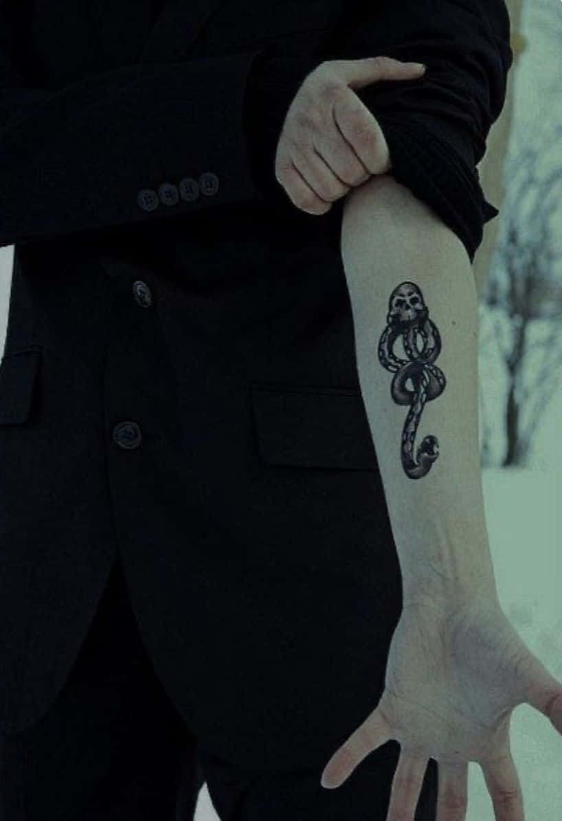Death Eaters 桌布 壁紙 — 一名身穿黑色西裝的人，在雪景背景下展示前臂上蛇纏繞骷髏的刺青。