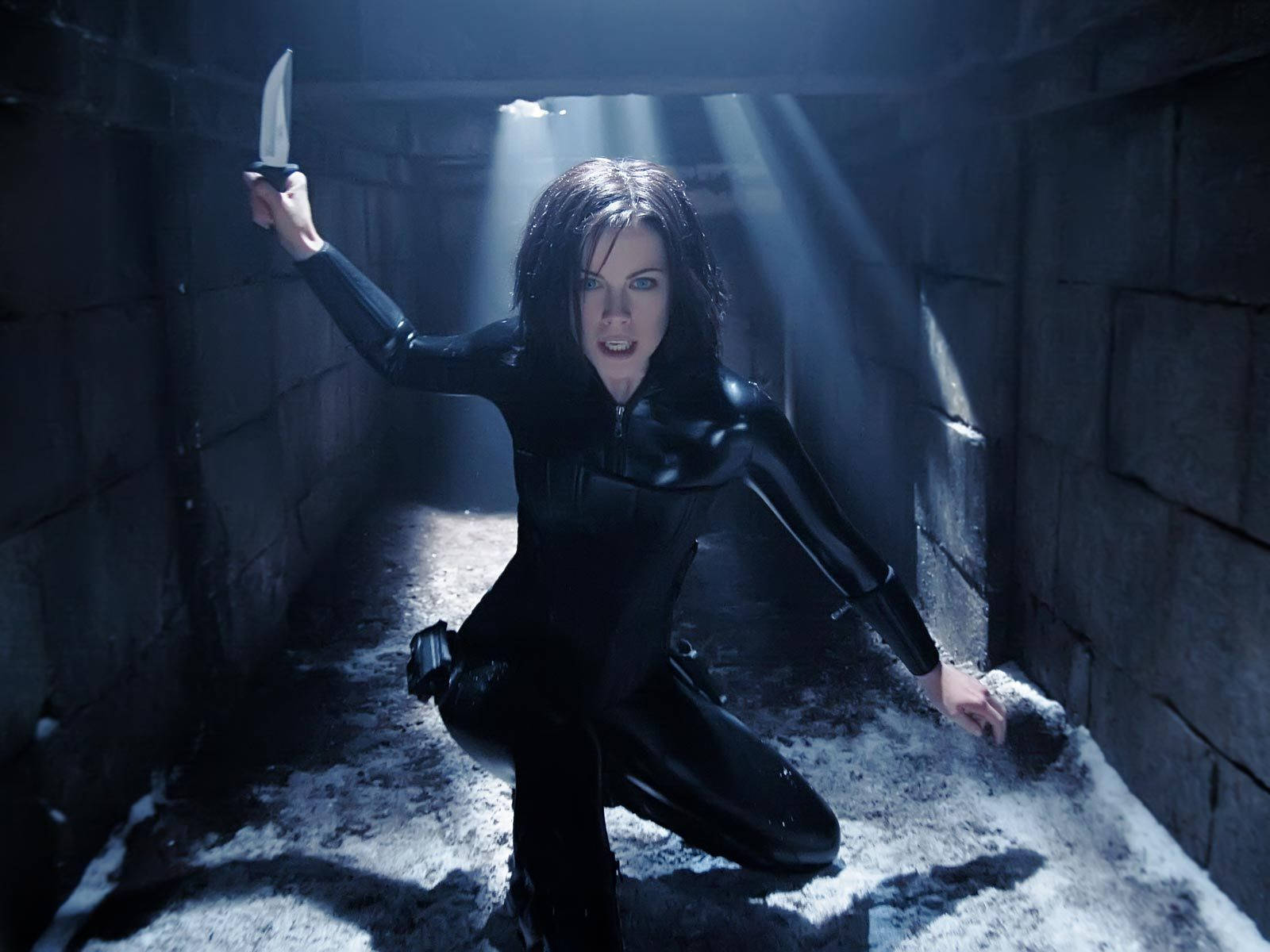 Kate Beckinsale Underworld 桌布 壁紙 — 一名身穿黑色西裝的女子蹲在石隧道中，手持一把刀，光束從上方刺穿黑暗。
