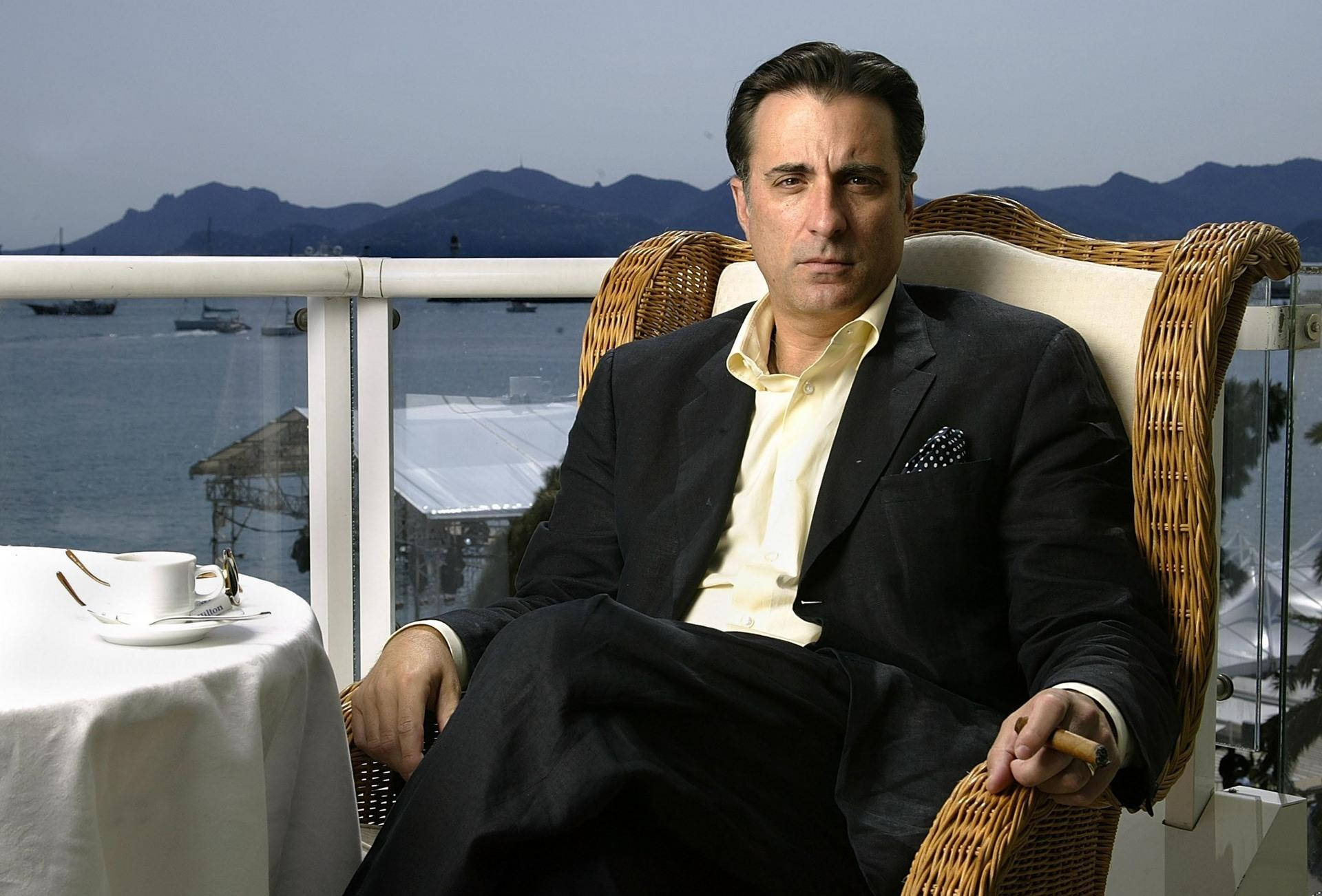 Andy Garcia 桌布 壁紙 — 一名身穿黑色西裝的男子，在一個俯瞰港口的陽台上，坐在藤椅上，港口有船隻和遠山，天空朦朧。