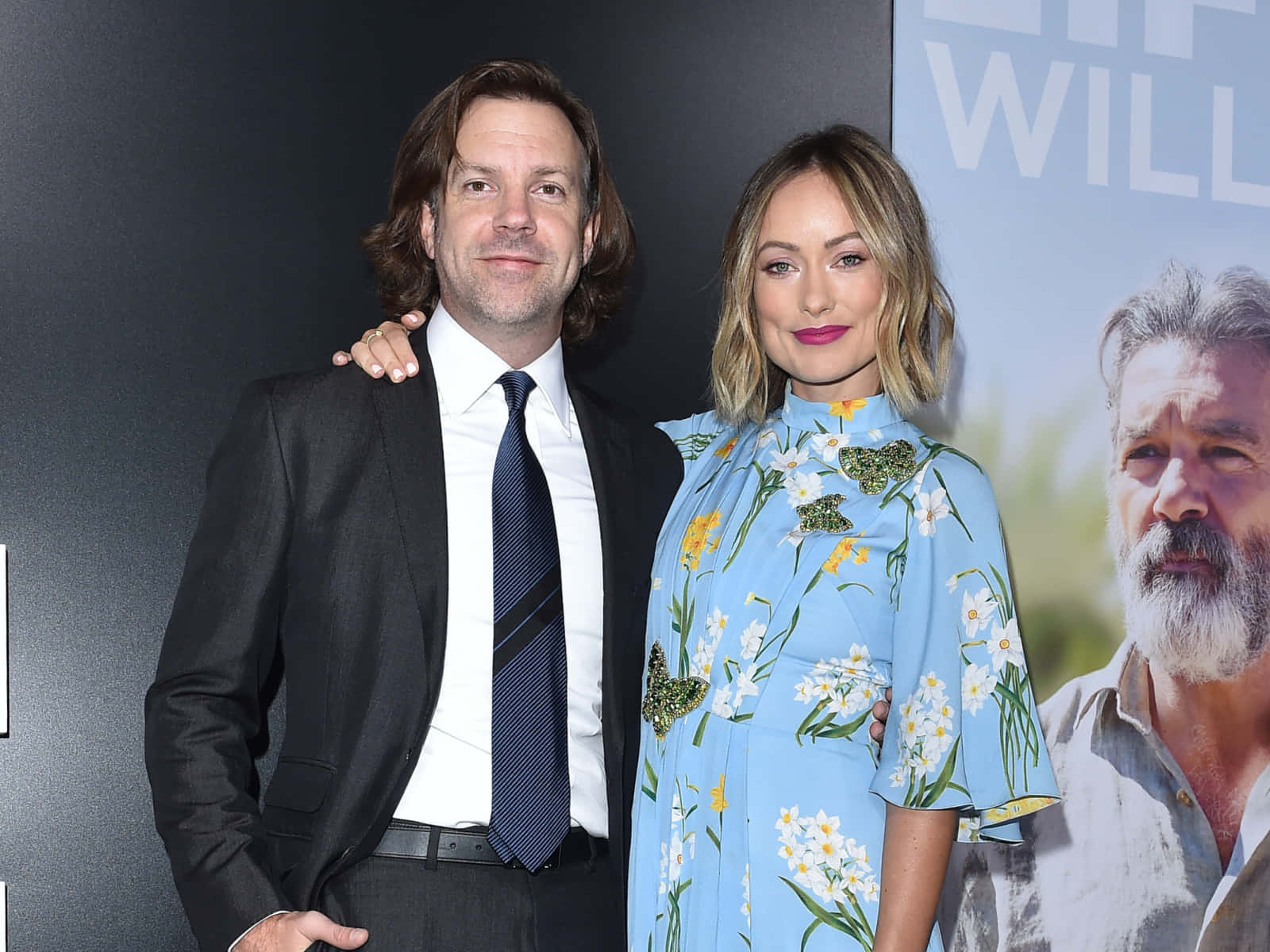 jason sudeikis 桌布 壁紙 — 一名身穿黑色西裝的男子和一名身穿花卉藍色連衣裙的女子在電影首映活動中一起擺姿勢。